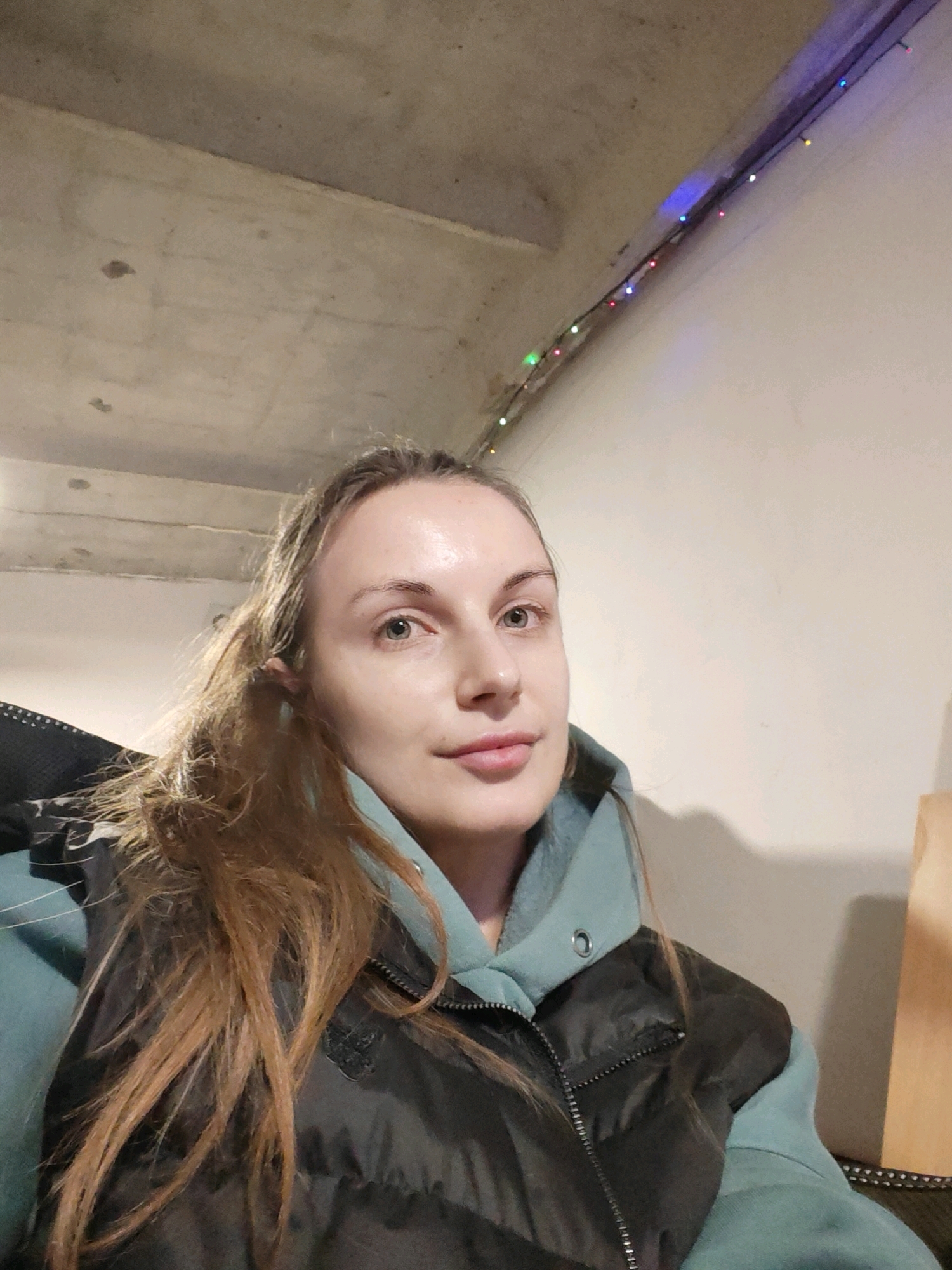 A woman with long, light brown hair is taking a selfie indoors. She is wearing a teal hooded sweatshirt and a dark puffy vest. The background consists of a concrete ceiling and a lightly colored wall with a string of colorful lights. The setting appears to be an indoor space, possibly a basement or unfinished room. There is no indication of the specific location within the city of Chaikovsky, Russia.