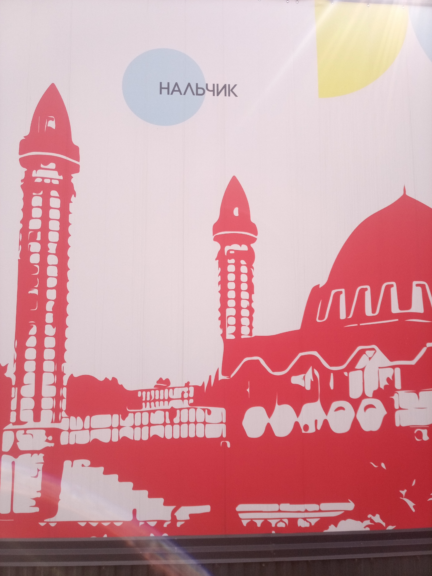 The image is a graphic representation of the city of Nalchik, Russia, featuring a stylized depiction of buildings and cultural elements. A prominent element is the text "НАЛЬЧИК" (Nalchik) written in dark grey, appearing within a light blue circular shape. To the left of the text, a red silhouette of a tall, slender tower, likely a minaret, is visible. To the right of the text, there is a red silhouette of a domed building, characteristic of a mosque or similar architectural style, with intricate white detailing on its facade. The foreground is dominated by a red silhouette of a cityscape, which includes smaller buildings, arches, and what appears to be a set of stairs. The background is a plain white surface, with abstract yellow and light blue circular shapes in the upper right corner, possibly representing abstract artistic elements or sunlight. The overall style is modern and minimalist, using bold colors and outlines to create a recognizable image of the city.