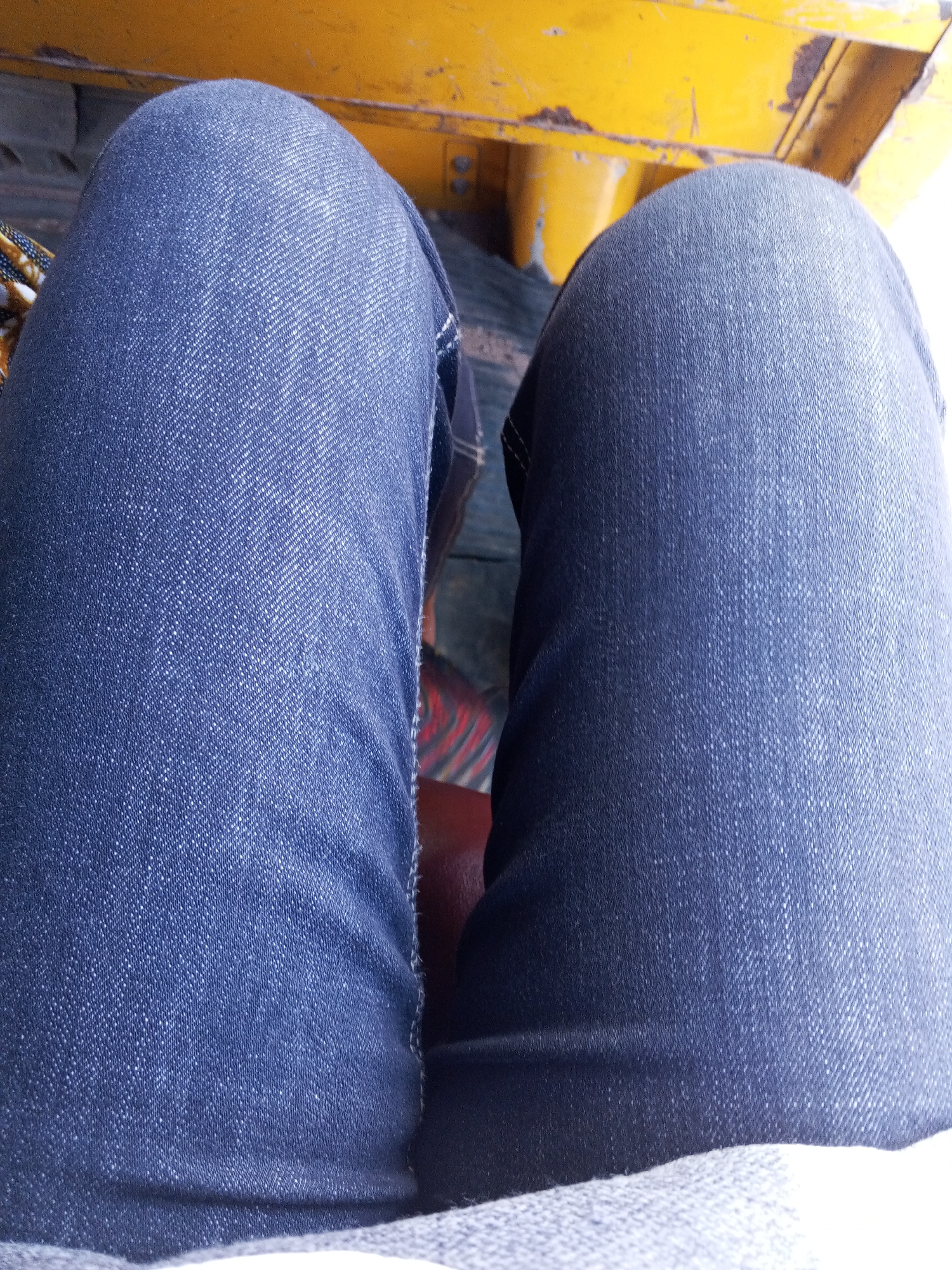 The image shows a close-up view of a person's legs from the knees down, both covered in blue denim jeans. The legs are positioned in a slightly spread stance. In the background, a bright yellow surface with some dark, possibly rusted, metal accents is visible, suggesting the person is seated in a vehicle or a similar structure. The overall lighting is bright, indicating an outdoor or well-lit indoor environment. No specific landmarks or features definitively place the scene in Jos, Nigeria, but the description adheres to the forensic, matter-of-fact tone requested.
