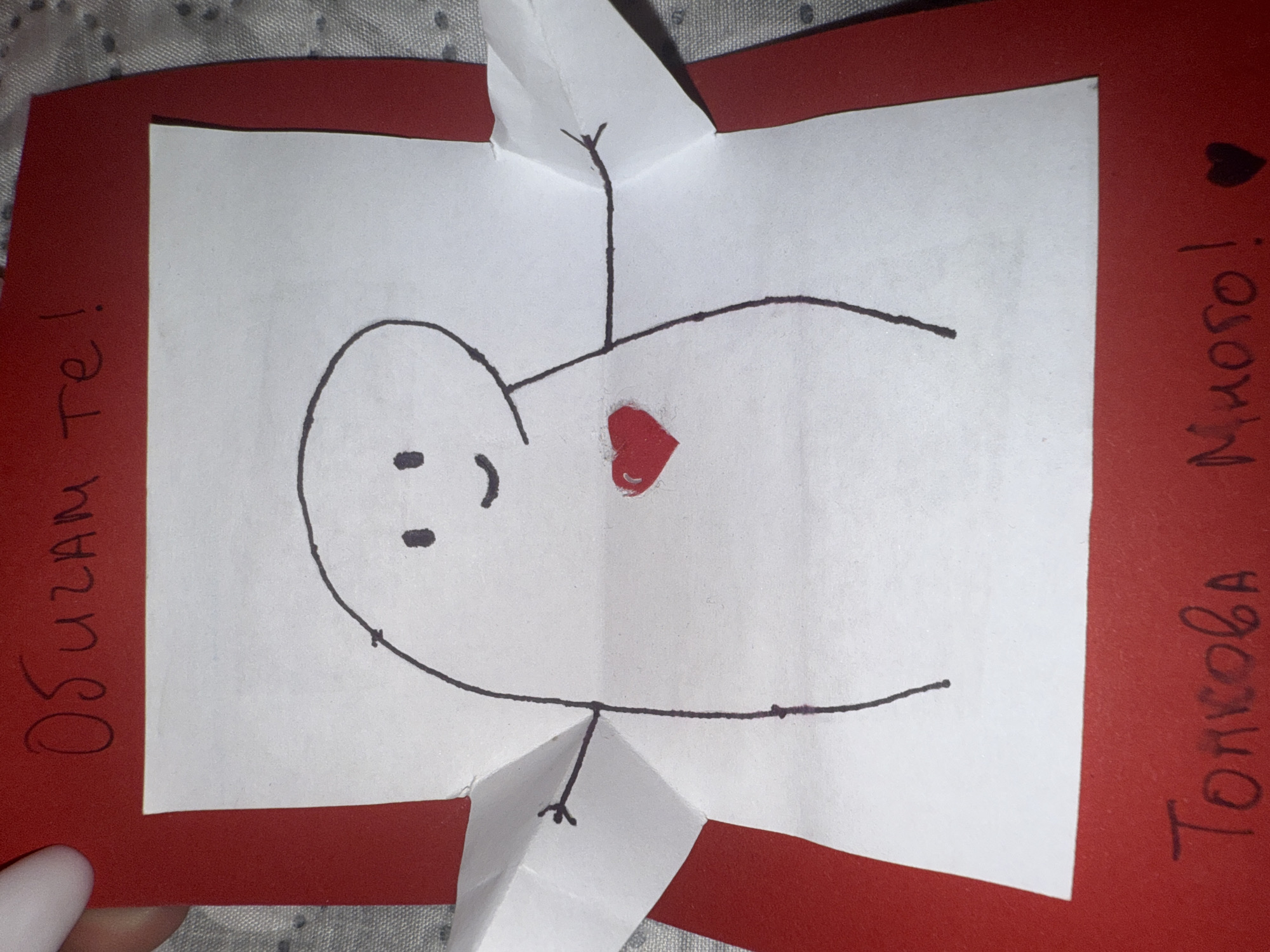 The image shows a handmade pop-up greeting card, crafted from red cardstock and a white paper insert. A simplified, childlike figure is drawn in black outline on the white paper, featuring an oval head with two dot eyes and a curved mouth. A bright red heart is prominently displayed on the figure's chest. The figure's limbs are part of the paper folds that create the pop-up effect.
Vertical text written in black marker is visible on the red cardstock. On the left side, it reads "Обичам те!" (Obicham te!), which translates from Bulgarian to "I love you!". On the right side, the text says "Толкова Много!" (Tolkova Mnogo!), meaning "So much!". A small black heart symbol is drawn above the word "Много!" on the right.
The card is placed on a light-colored, textured surface, possibly a patterned fabric, suggesting an indoor setting. The overall presentation conveys a heartfelt and affectionate message. The Bulgarian language used in the text aligns with the provided context of Varna, Bulgaria.