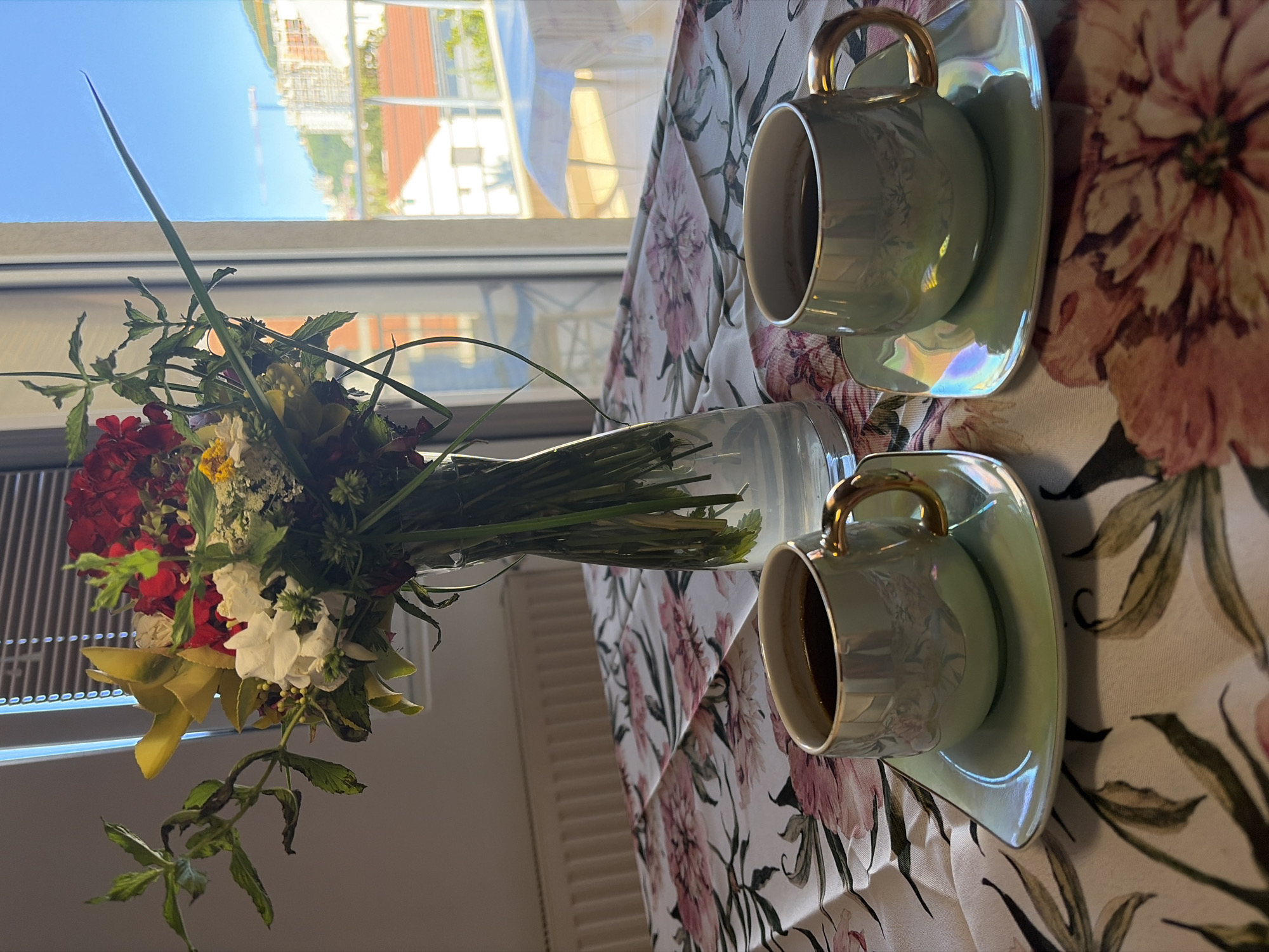 A still life photograph shows a flower arrangement, a clear glass vase with water, and two cups of coffee with saucers on a floral tablecloth. The background shows a window with a view of buildings and the sky, suggesting the scene is inside an apartment or residence. The location may be Budva, Montenegro.