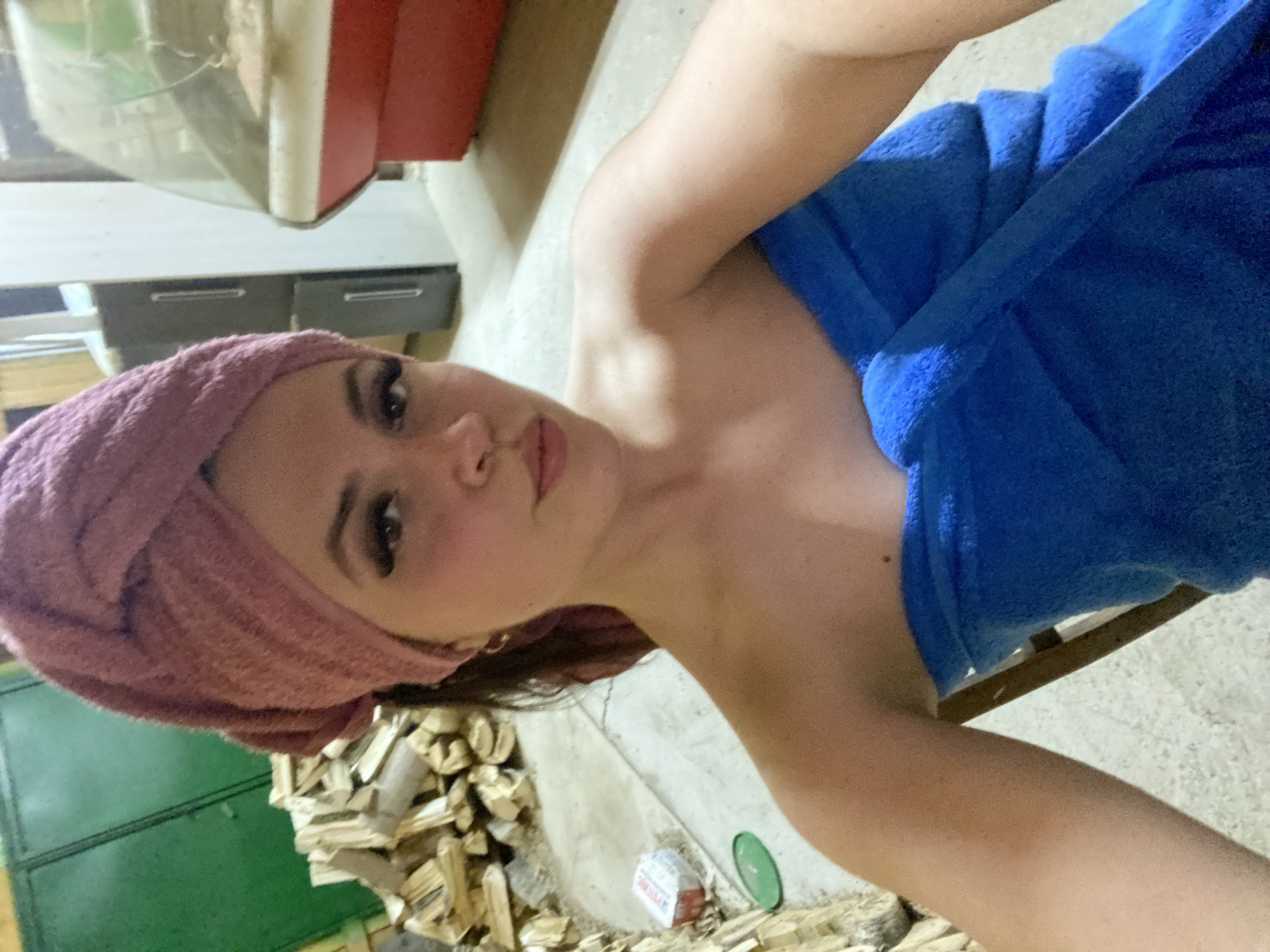 A selfie photograph depicts a young woman with long brown hair partially wrapped in a pink towel, who is looking directly at the camera. She is partially covered by a blue towel and is positioned indoors. Background elements include a pile of logs, and what appears to be a stove, with a green door in the lower left corner. The location is Zhigalki, Russia.