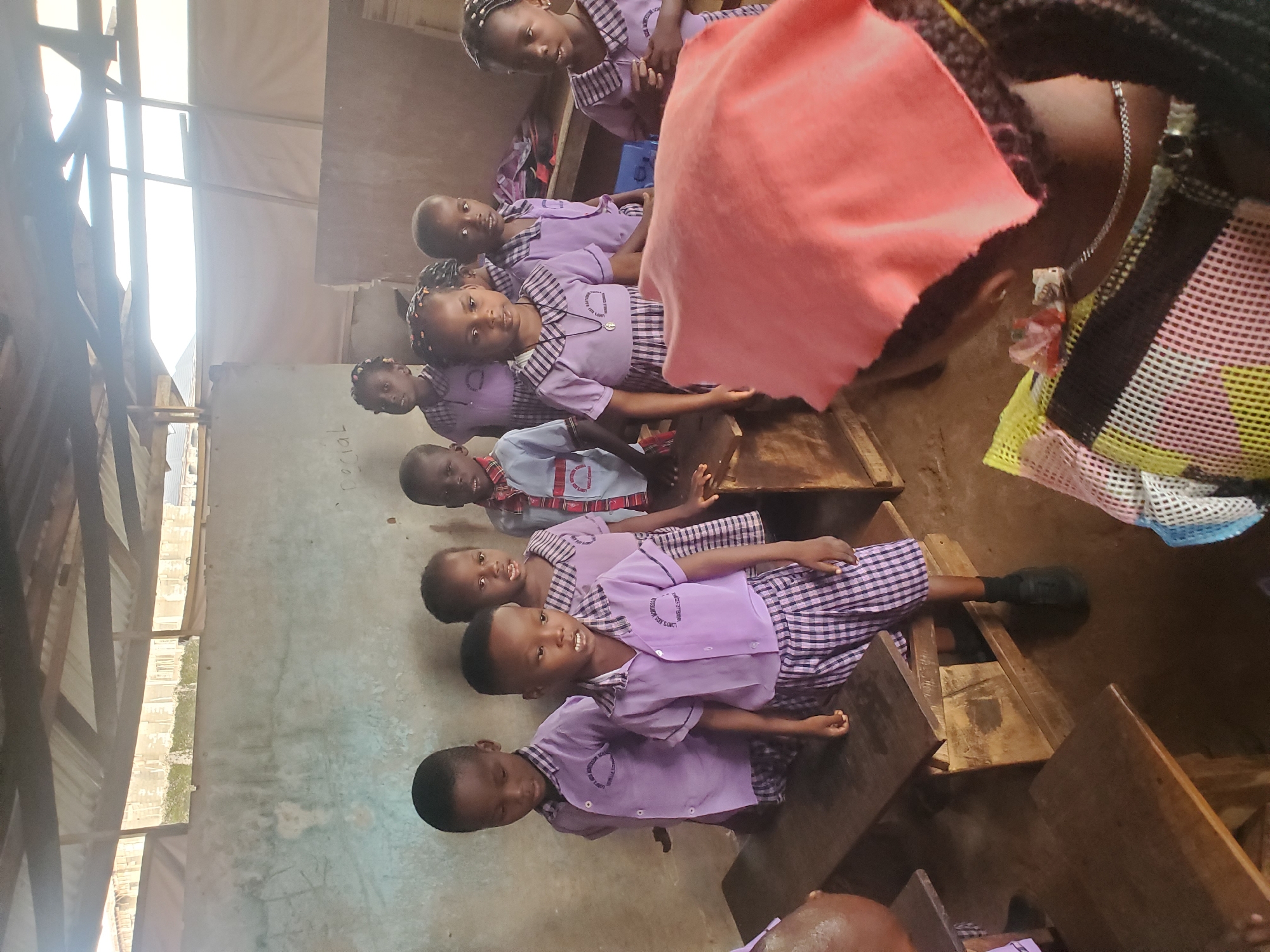 The scene is an interior classroom in Ukwu-Abwa, Nigeria. A group of students, primarily children, are seated at wooden desks. They are wearing purple school uniforms. A woman, partially visible in the foreground, is wearing a mesh top with a chain. The classroom appears to be in a building with a tin roof. The walls are stained and bare, except for a blackboard on the wall. The children are looking towards the viewer.