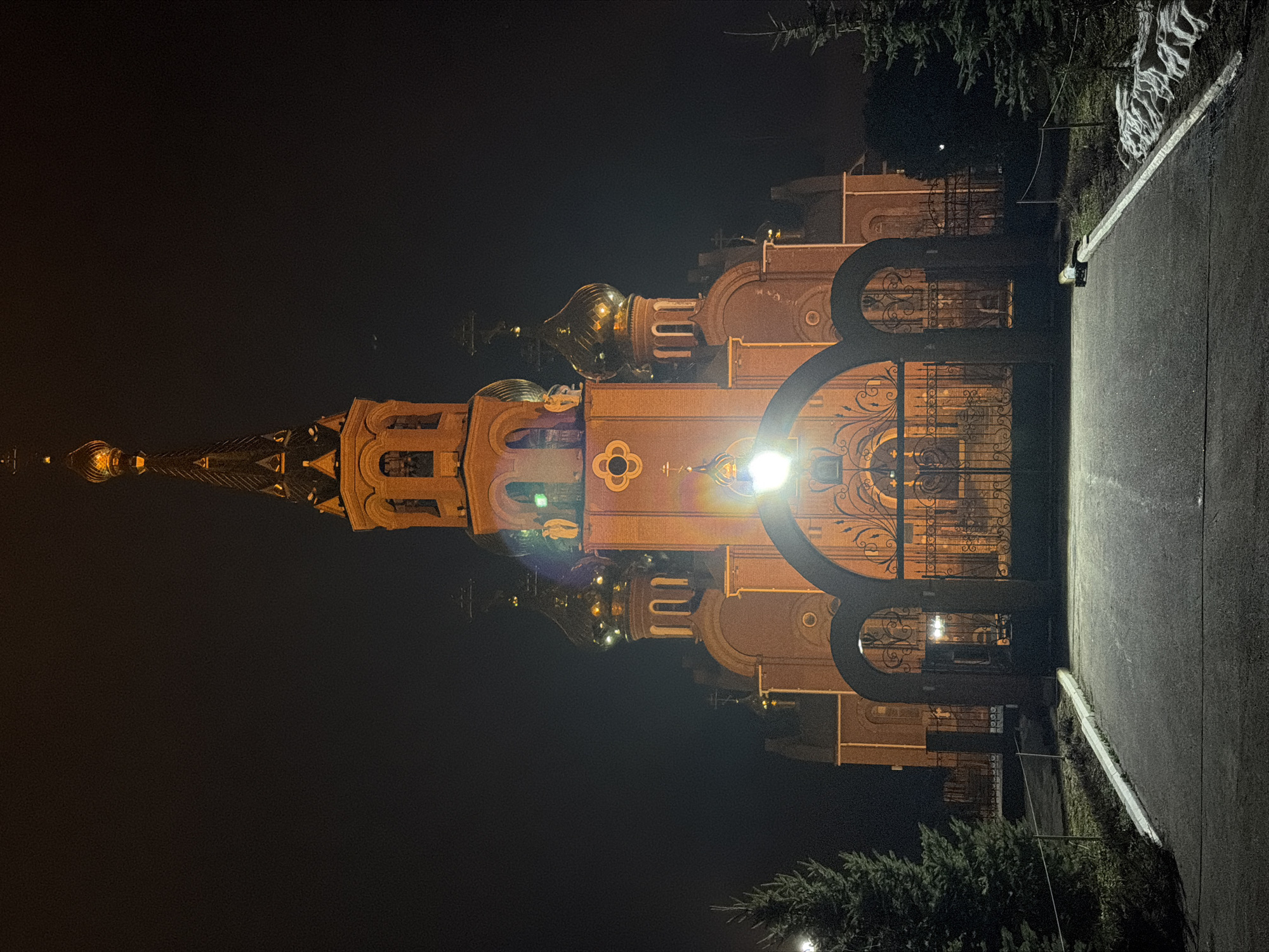 The image depicts the Orthodox Church of St. George the Victorious in Chaikovsky, Russia, at night. The church is illuminated, highlighting its architectural details, including golden domes and an intricate facade. A dark wrought-iron gate stands in the foreground, with a pathway leading towards the church. The sky is dark, and surrounding vegetation is visible in the lower corners of the frame. A street light is shining directly in front of the church, causing a light flare.