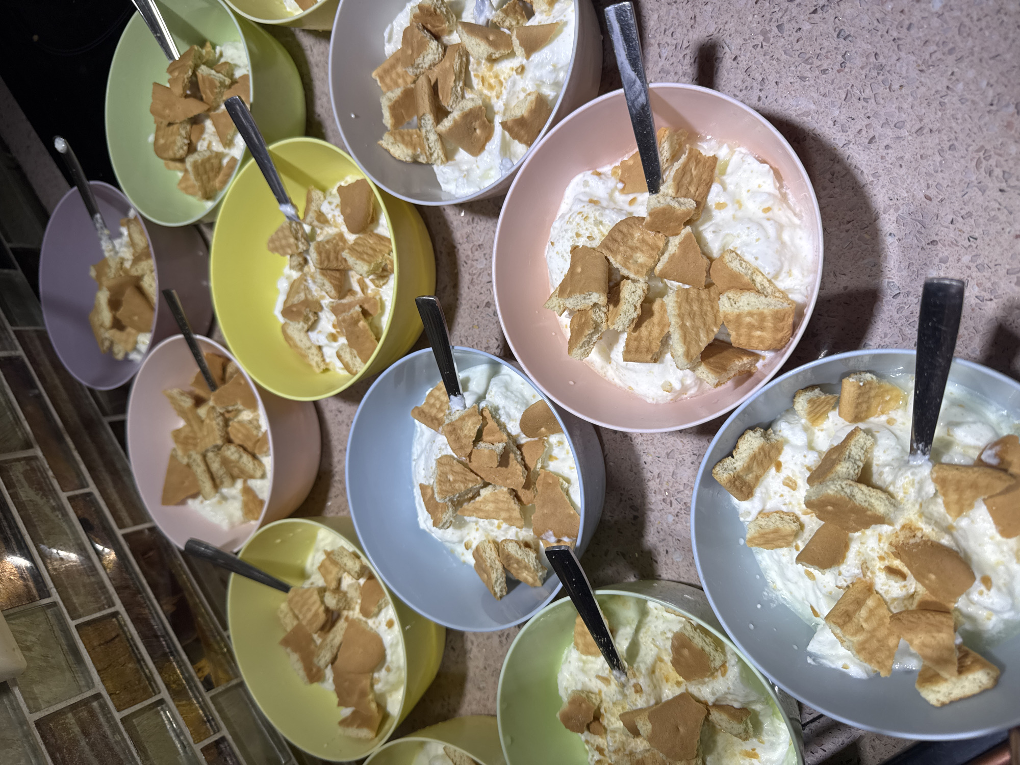 The image shows multiple colorful bowls containing a dessert, likely banana pudding or a similar treat, consisting of a creamy white base topped with pieces of a golden-brown wafer-like biscuit. Each bowl contains a silver spoon. The bowls are arranged on a speckled countertop. There is no evidence to suggest this image was taken in Sofia, Bulgaria, and no people are visible.