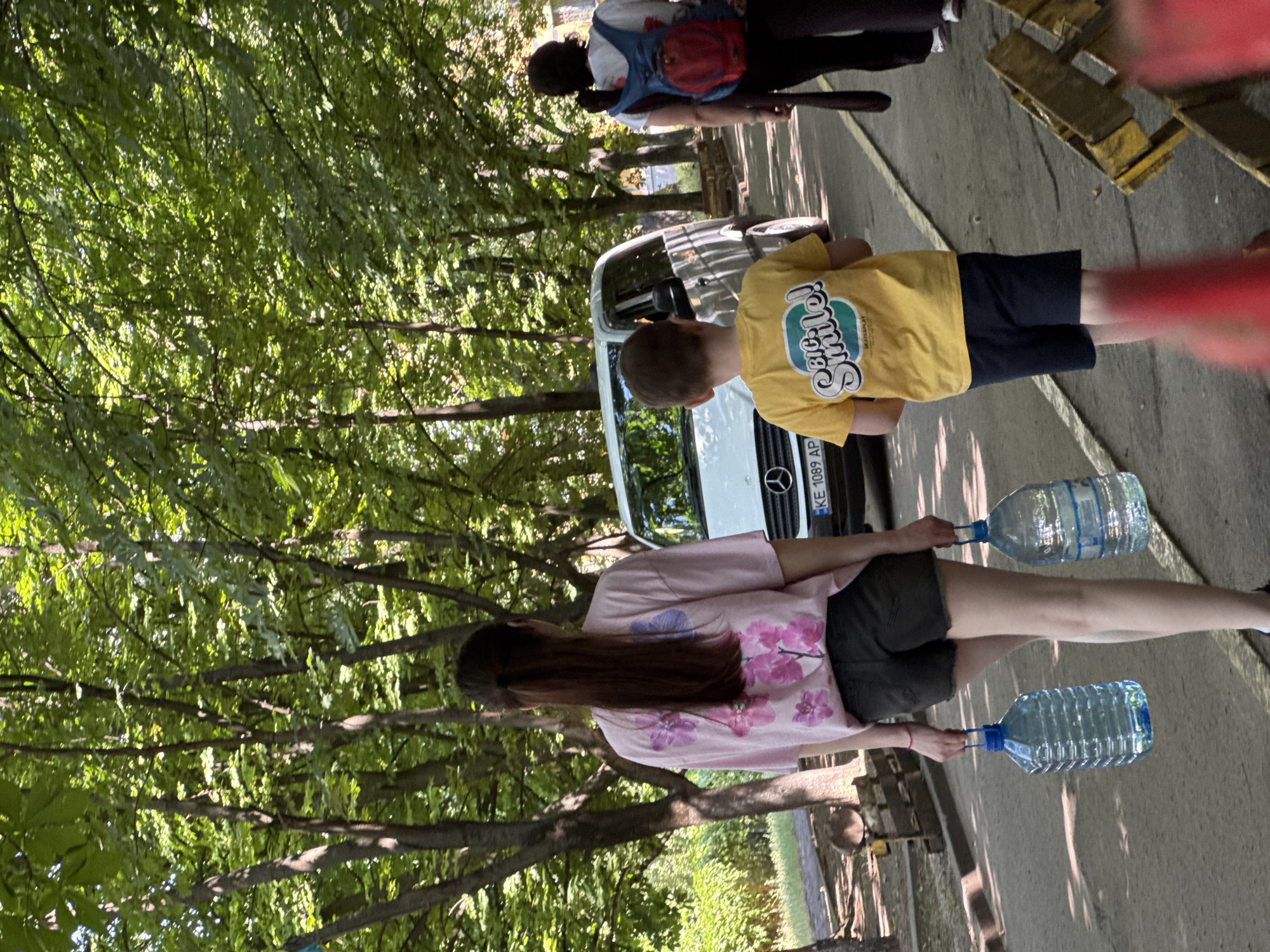 The image shows two people carrying large plastic bottles of water down a paved path lined with trees. The person in the foreground is a woman wearing a pink t-shirt with a floral print and dark shorts. She is carrying two full bottles of water, one in each hand. Her long, dark hair is visible. Behind her, a boy in a yellow t-shirt with the text "Smile! Be Cool!" and dark shorts is also carrying a bottle of water. He appears to be walking slightly ahead of the woman. In the background, a white van is parked, and another person, possibly a woman, wearing a backpack, is visible near the van. The scene is outdoors, with dappled sunlight suggesting it is daytime. The trees are lush and green, indicating a warm season. The setting appears to be a park or a similar outdoor recreational area. The overall mood seems neutral, possibly depicting an everyday task or an activity related to the van. There is visible text on the yellow t-shirt of the boy and on the license plate of the van, which reads "KE 1089 AP". The overall context suggests a summer day in Kryvyi Rih, Ukraine.