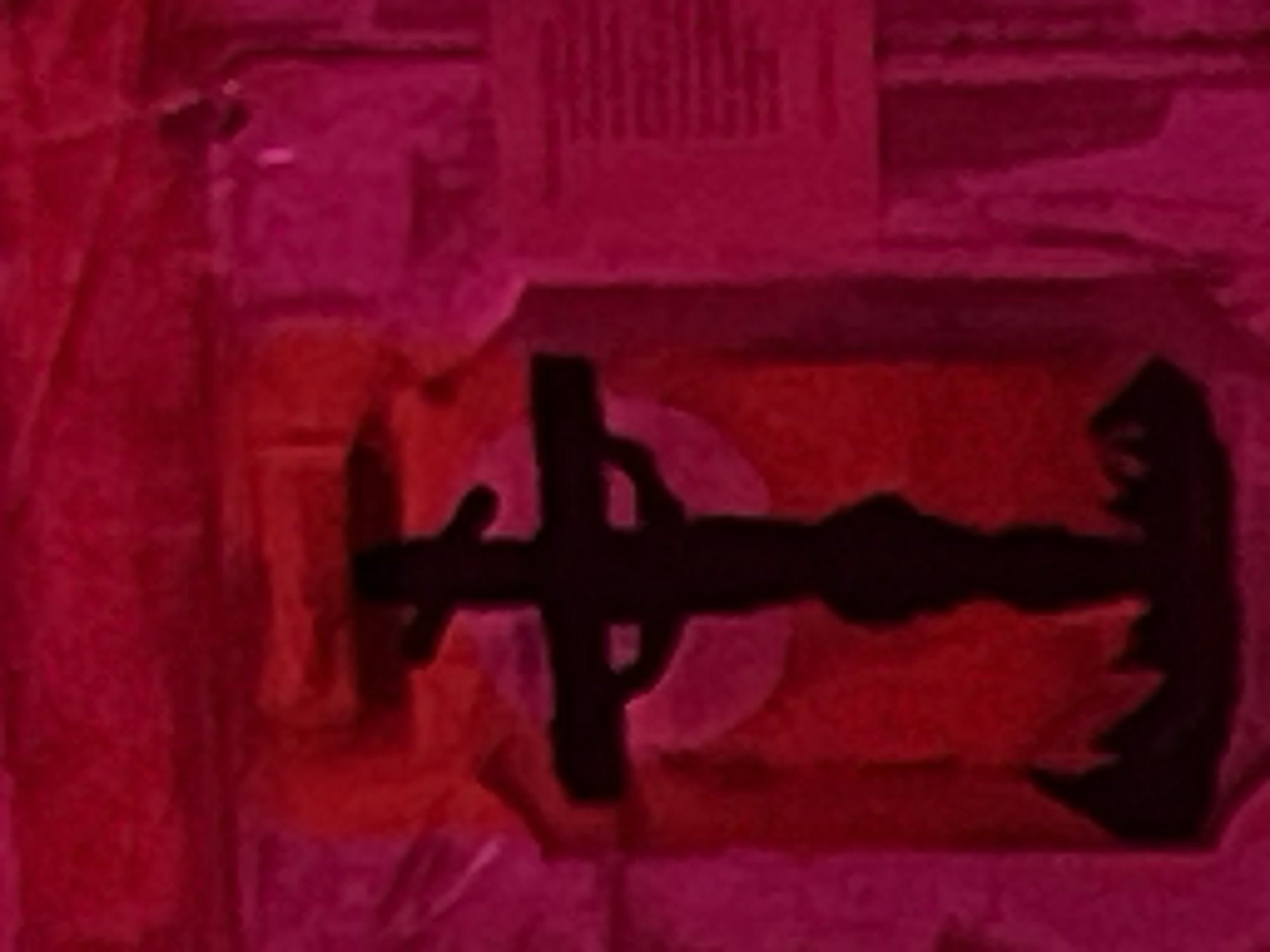 The image displays a dimly lit, red-toned scene in Debelt, Bulgaria, featuring a prominent, dark silhouette of a crucifix. The crucifix, centered within a recessed niche, depicts a human figure on a cross, with outstretched arms and a body hanging downwards. The base of the cross rests on a dark, jagged, irregular form, possibly representing rocky terrain or a decorative element. Behind the upper portion of the crucified figure, a lighter red, circular glow is visible within the niche. To the right of the niche, a pale, rectangular plaque or sign is affixed to the wall, displaying multiple lines of indistinct text. The surrounding environment consists of a textured wall bathed in a deep red and magenta light. No individuals are present in the frame.