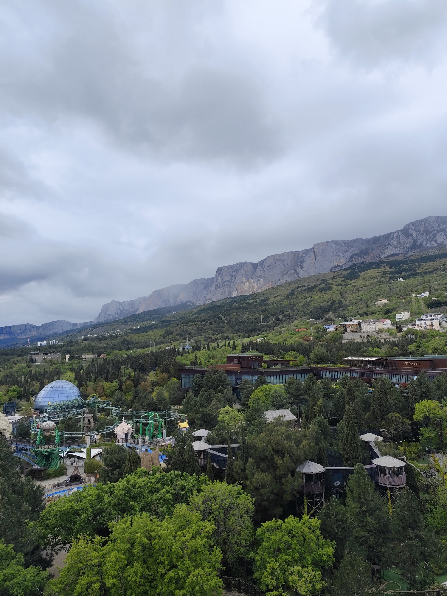 The image shows an overview of the city of Симеизский поселковый совет, Ukraine, under a cloudy sky. In the middle ground, a modern, horizontally oriented building with dark windows, possibly a hotel or resort, is visible. The building is identified by signs on its facade.
In the foreground, there is a dense area of trees, among which parts of what appears to be an amusement park are visible. This includes a distinctive blue dome structure and sections of a roller coaster. Several wooden towers are scattered around.
In the background, rugged mountains rise above the cityscape, partially obscured by haze or low-hanging clouds.
The scene suggests a location where tourist attractions are integrated into the natural environment.