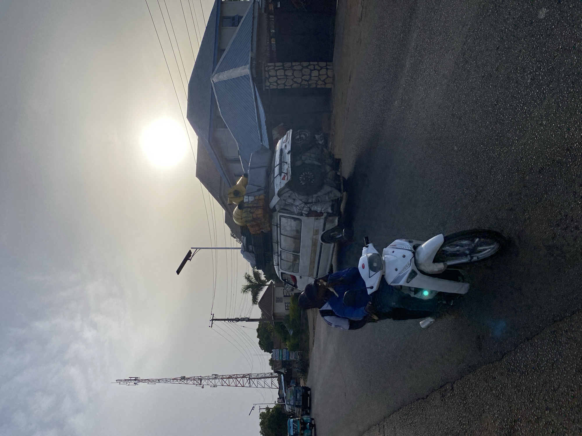 A street scene in Jos, Nigeria, shows a man on a white motorbike parked on an asphalt road. Next to him is a white van with items strapped to the roof. Buildings are on the left and other vehicles are parked in the distance. A tall communications tower is also visible. The sky is bright and overcast.