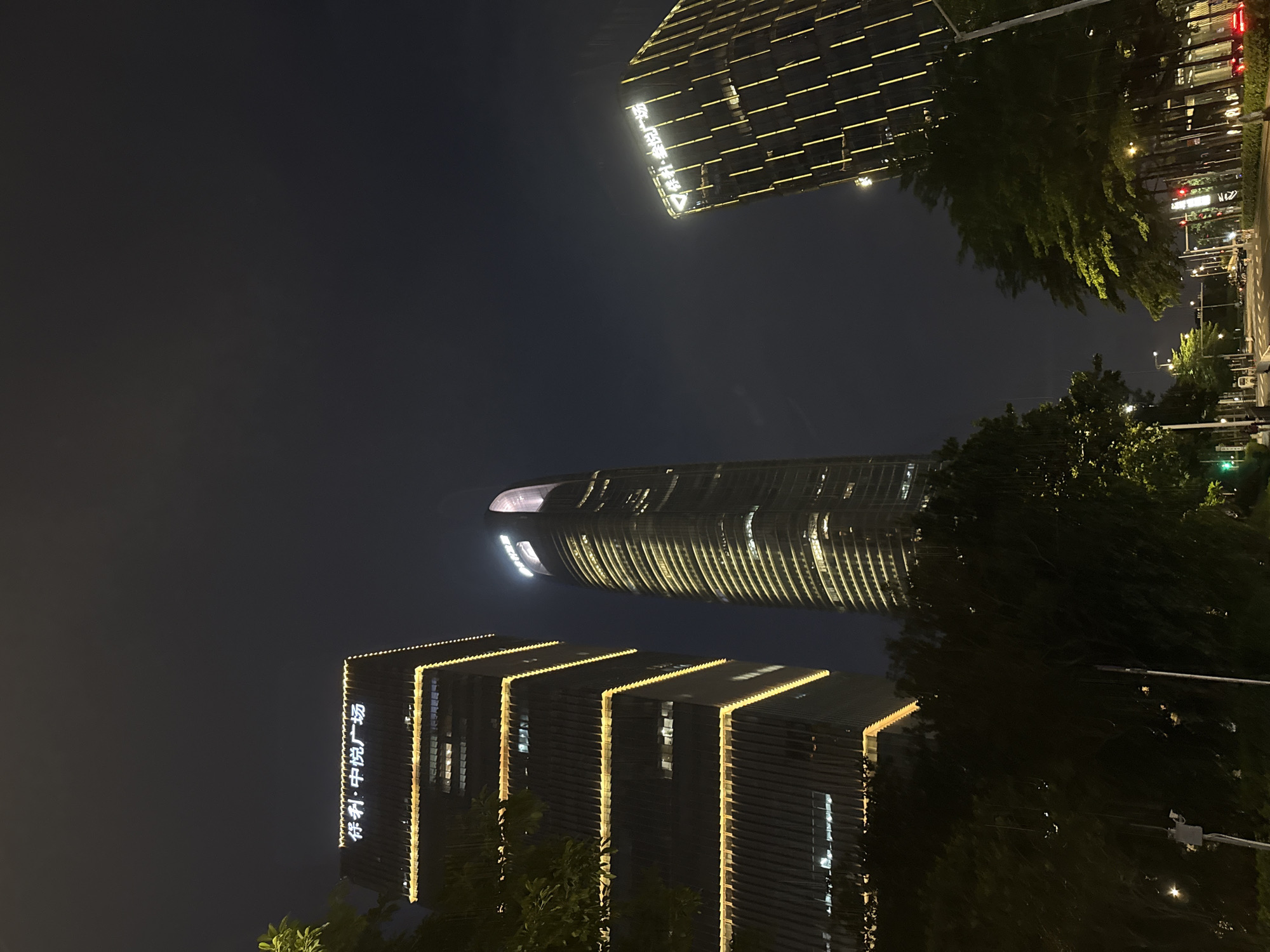 This nighttime urban landscape captures three brightly illuminated high-rise buildings from a street-level perspective in Haizhu District, Guangzhou, China.
On the left, a multi-story, rectangular building with a stepped facade is prominently lit by horizontal yellow light strips at each setback. The building's upper section displays luminous white Chinese characters reading "保利・中悦广场" (Bǎolì・Zhōngyuè Guǎngchǎng), identifying it as Poly Zhongyue Plaza.
In the center, a very tall, slender, cylindrical or elliptical skyscraper dominates the skyline. This building is extensively illuminated with both vertical and horizontal yellow light lines, accentuating its height and curved form. Its distinctive rounded top glows with a soft white-purple light, and faint Chinese characters are visible near its apex, though not clearly decipherable. This building is characteristic of the Guangzhou International Finance Center (GZIFC).
On the right, another high-rise building with a rectangular profile is visible, illuminated by vertical yellow light strips along its facade. The upper part of this building features luminous white Chinese characters reading "越秀・星汇文渊" (Yuèxiù・Xīnghuì Wényuān), identifying it as Yuexiu Xinghui Wenyuan.
The foreground is filled with dense, dark green foliage from numerous trees and bushes, obscuring the lower portions of the buildings. A multi-lane asphalt road is visible towards the bottom right, bordered by more trees and streetlights emitting a warm glow. Traffic lights are visible in the distance, one showing red and another green. A single person is faintly discernible walking on a sidewalk on the far left. The sky is dark and appears to be overcast, with some atmospheric haze reflecting the city lights.