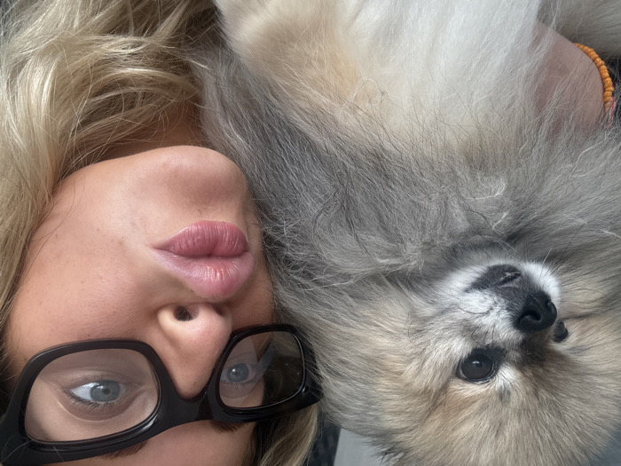 A close-up image features an adult female interacting with a Pomeranian dog in an indeterminate indoor setting. The female, positioned on the right, has blonde hair and wears black-framed glasses. Her lips are pursed in a "kiss" gesture, and her visible left eye is blue. A portion of her hand, adorned with an orange beaded bracelet, is visible, indicating she is holding the dog. The Pomeranian, occupying the left and central frame, exhibits tan and greyish-white fur, dark eyes, and a black nose. Its head is turned slightly to the left, away from the camera. The location in Wattrelos, France, is not discernible from the visual information provided within the image.