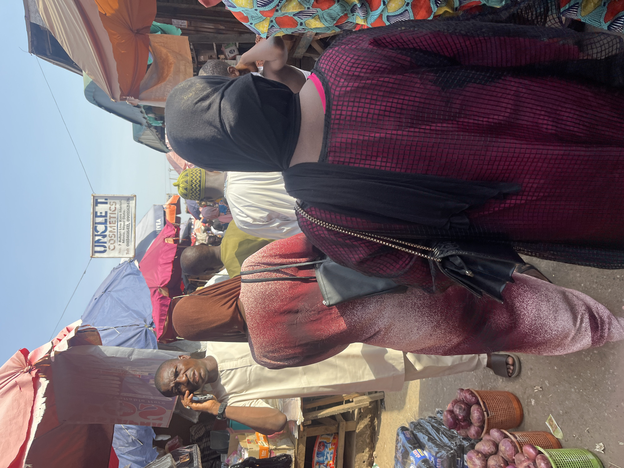 The image shows a busy market scene in Minna, Nigeria. Multiple individuals are present, including one man talking on a mobile phone under colorful umbrellas. Some individuals are wearing traditional clothing. The foreground contains baskets filled with red onions next to packaged goods. In the background, signage for "Uncle T. Cosmetics" is visible, providing a business reference point within the Kure Market.