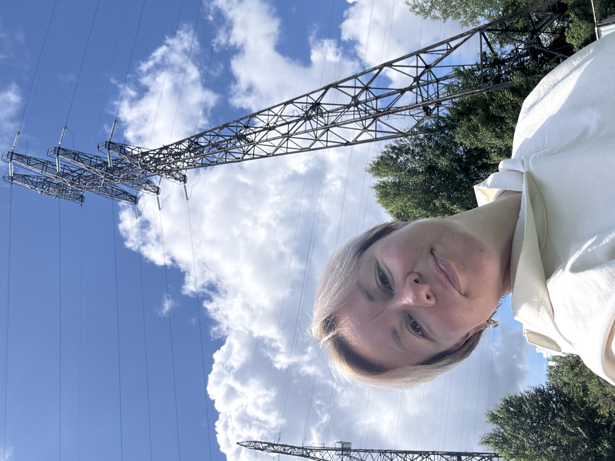 The image is a selfie taken outdoors. A person with short, light-colored hair is positioned in the foreground, partially obscuring the view. The background features a clear blue sky with puffy white clouds, and power lines supported by metal towers. Green trees are also visible. The setting appears to be near an industrial area, possibly on the outskirts of a city. There is no additional evidence to confirm this image was taken in Chaikovsky, Russia.