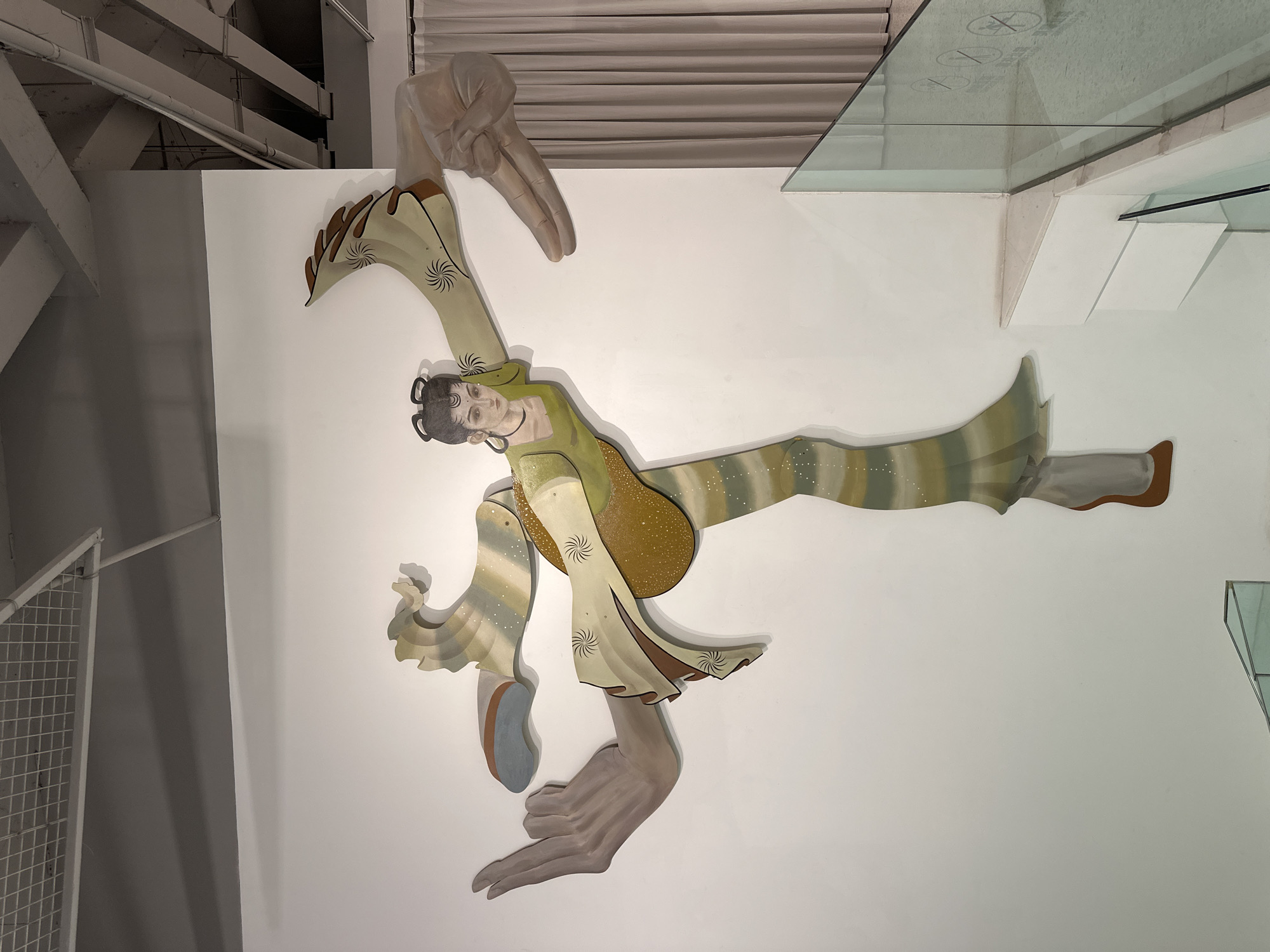 A photograph depicts an abstract mural on a white wall in an interior space. The mural is comprised of painted wooden shapes that together form a figure lying diagonally across the frame. The figure has a human face and torso, but its limbs are extended and stylized, ending in two large, painted hands. The hands appear to be supporting or manipulating the figure. The figure is dressed in a green and tan outfit with brown accents. The overall aesthetic is surreal and art deco-inspired.
The background consists of a white wall, with the top right corner showing the edge of a glass enclosure and a beige-colored curtain or blind. The top left corner reveals exposed structural elements of the building, including beams and pipes. The bottom left corner has a glimpse of a metal grate.
The image provides no evidence that the location is in the city of Banan, China.