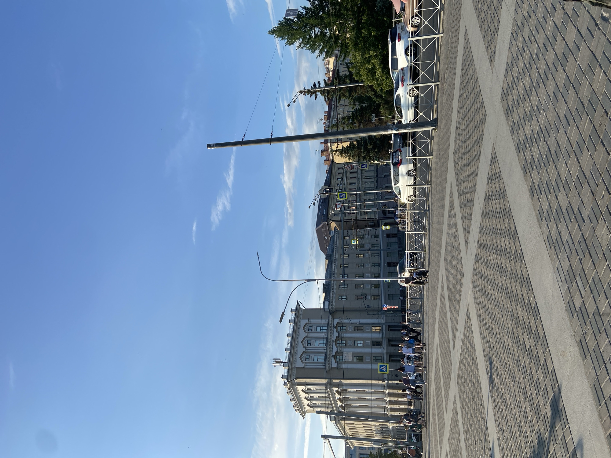 The image captures a street scene in Kazan, Russia. A wide pedestrian walkway paved with gray brick tiles is the focal point. Buildings line the street, including a multi-story structure with intricate facade details and classical architectural elements. People are present, some walking along the walkway. Cars are parked along the side of the street, separated from the walkway by a metal railing. Tall streetlights and utility poles punctuate the scene, and a green tree adds a touch of nature. The sky is a clear, bright blue with some thin clouds.