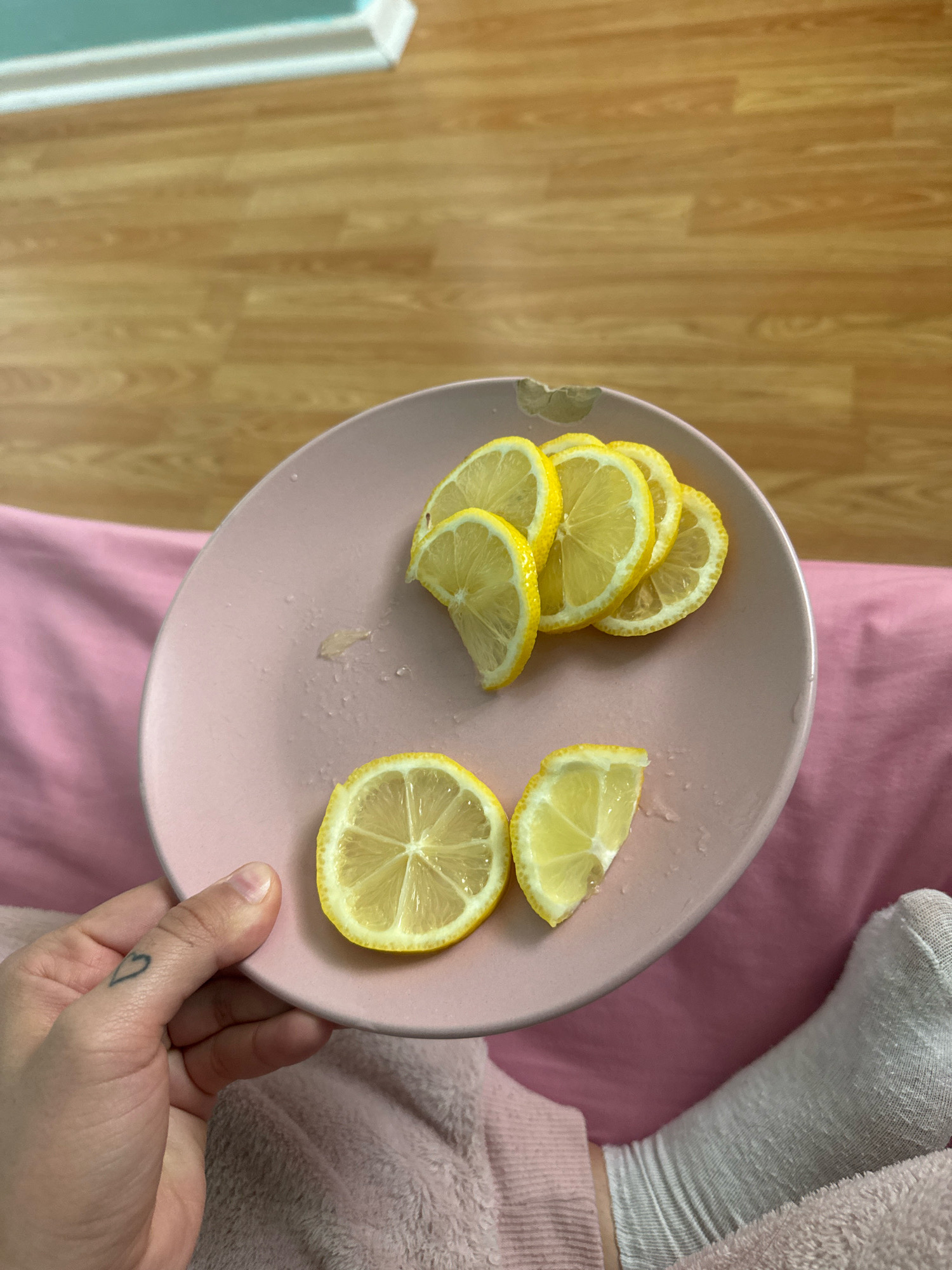 A person's hand with a small heart tattoo on the index finger is holding a pale pink plate. The plate contains several slices of lemon, some of which are partially overlapping. A small piece of lemon rind and some juice droplets are visible on the plate. The plate is positioned over a pink fabric, likely a blanket or clothing. In the background, a wooden floor is visible, along with a white baseboard and a turquoise wall. A person's socked foot is also present in the lower right portion of the image. The scene appears to be indoors.