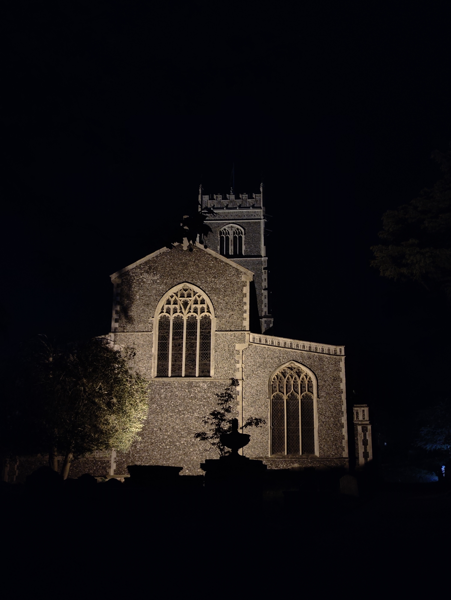 The image depicts a church at night. The building is made of stone, with two large arched windows visible. A tower rises from the back of the church. The church appears to be illuminated by spotlights, creating a stark contrast between the bright facade and the dark background. Trees are seen on either side of the building. The ground in front of the church is dark, possibly a graveyard. The location is East Suffolk, United Kingdom.