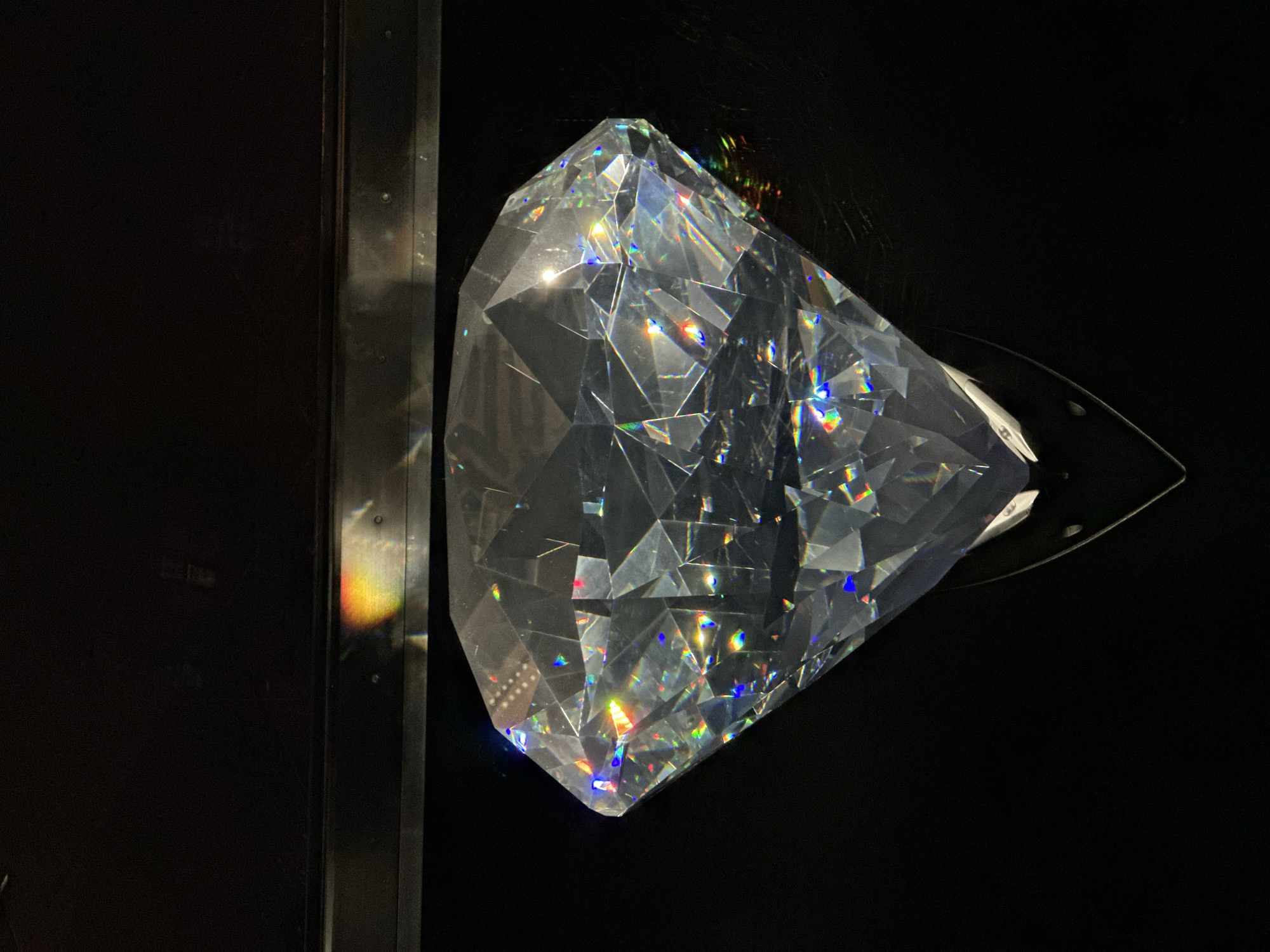 The image shows a large, faceted, clear crystal object resembling a diamond. The crystal is positioned centrally against a black background. Light is refracting through the crystal, creating a spectrum of rainbow colors on its facets and in the surrounding area. The crystal appears to be displayed in a dark environment, possibly a display case or a darkened room, given the reflections seen at the top of the image. The specific location in Wattens, Austria, is not discernible from the image itself, but the presence of such a crystal would likely indicate a museum, gallery, or shop related to crystal production or display, given Wattens is known for Swarovski Crystal Worlds. The scene is static and focuses solely on the object and its interaction with light.