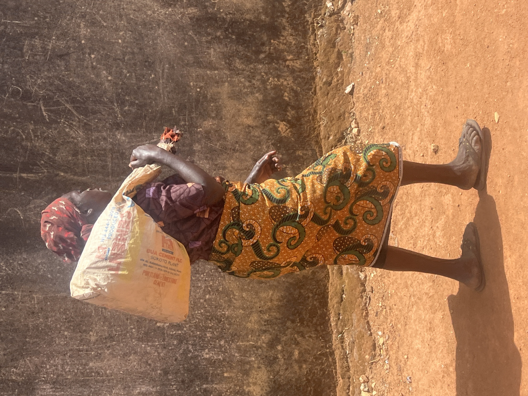 An outdoor, full-body shot depicts a person, likely a woman, carrying a large, heavy white sack on her head, walking along a dusty path in Gura Top, Nigeria. The image is captured in bright, direct sunlight, casting strong shadows on the reddish-brown ground, indicating midday or early afternoon. The weather appears clear and sunny.
The person is dressed in traditional attire, featuring a vibrant patterned skirt or wrapper with a predominantly orange, green, and brown swirl design, paired with a darker patterned top and a reddish headscarf. She wears simple dark sandals. Her arms are bent, assisting in steadying the load.
To her left, a dark, textured wall or surface, possibly made of mud or rough plaster, forms the background. The ground is a reddish-brown earth, dusty and uneven, with small stones scattered throughout.
The visible text on the large white sack provides specific details. It reads: "BUA CEMENT PLC", "SOKOTO PLANT", with an address of "KM 10, Kalambaina road, Sokoto, Sokoto State". Other visible text includes "H.S. 444-1:2018" and the product type, "PORTLAND LIMESTONE CEMENT". The brand name "BUA" is also partially visible. The scene conveys an everyday activity of transporting goods, highlighting the physical labor involved in the setting.