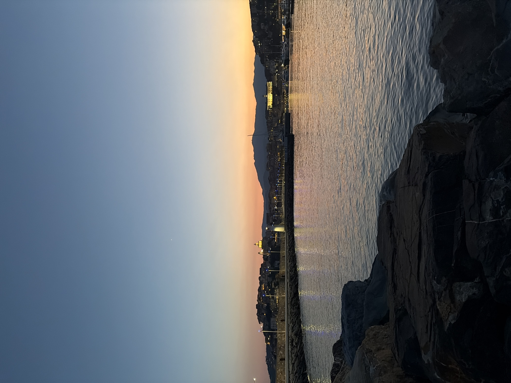 The image depicts the harbor of Imperia, Italy, during the twilight hours. In the foreground, dark, rugged rocks are visible, leading to the calm waters of the harbor. The water reflects the soft colors of the sunset, with hues of orange, pink, and blue blending in the sky.
The middle ground features a stone pier lined with streetlights that cast a warm glow on the water. Beyond the pier, the city of Imperia is illuminated by numerous lights, creating a picturesque silhouette against the darkening sky. A prominent church with a golden dome stands out on the hillside, a recognizable landmark of the city.
In the background, the gentle slope of the hills surrounds the harbor. The sky transitions from a clear, pale blue at the top to a warm gradient of colors near the horizon, indicating the end of the day. A few boats are moored in the harbor, their masts subtly visible against the illuminated cityscape. There are no people clearly visible in the frame. The overall atmosphere is serene and tranquil, capturing the beauty of a coastal Italian city at dusk.