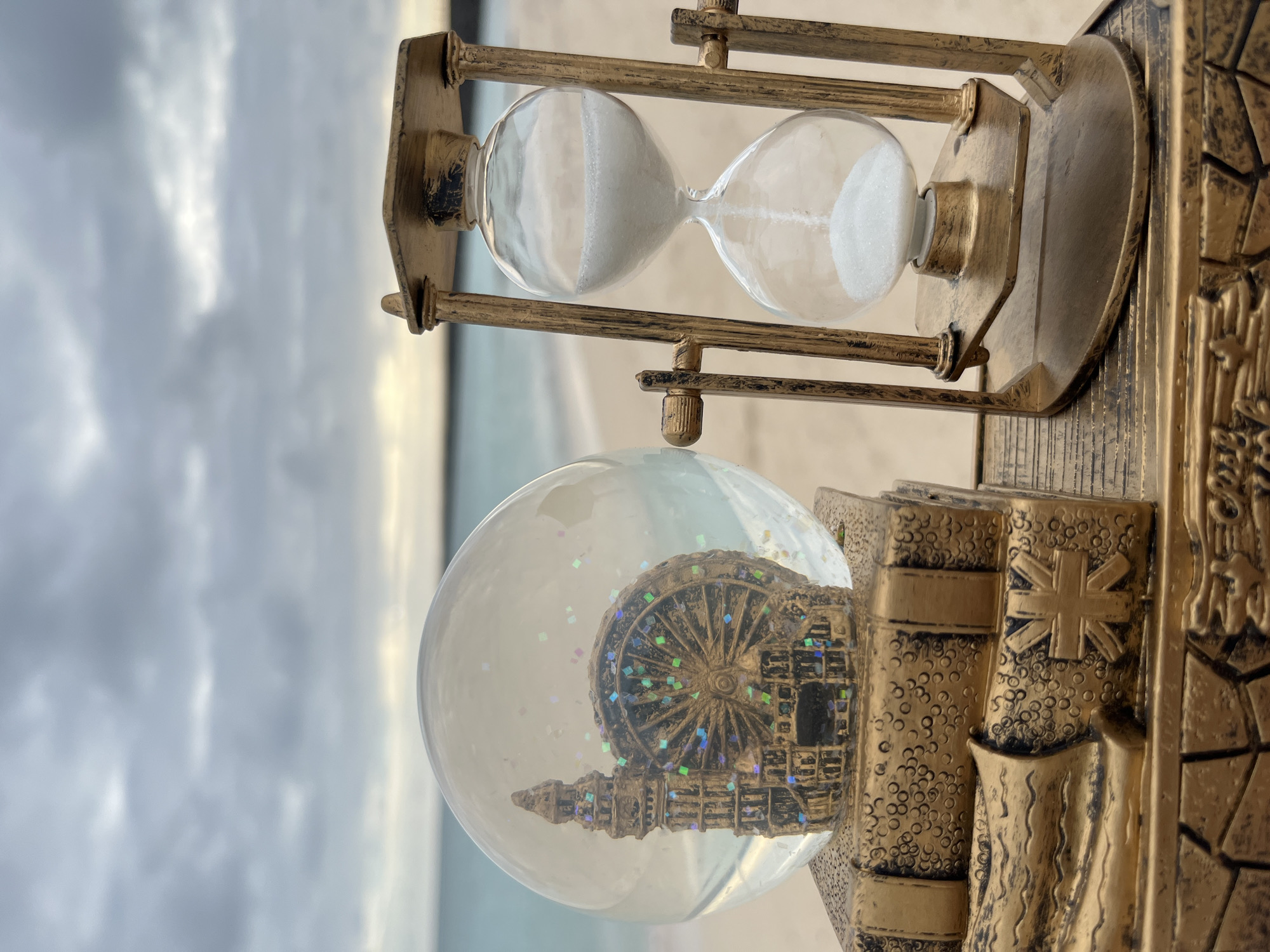 The image displays a close-up, low-angle shot of a snow globe and an hourglass on a beach. The snow globe, positioned on the left, contains a miniature representation of a Ferris wheel and a clock tower, possibly Big Ben, covered in glitter. It rests on a decorative base resembling stacked books. To the right of the snow globe is a golden hourglass with white sand flowing from the upper bulb to the lower one. Both the snow globe and the hourglass appear to be part of a larger, ornate golden decorative piece. The background features a blurred seascape with a calm ocean and a cloudy sky. The location is identified as Kuta, Indonesia, though this is not visually evident from the image itself. There are no people or animals visible in the scene, and no actions or interactions are depicted. The overall environment is a beach with sand and the ocean.