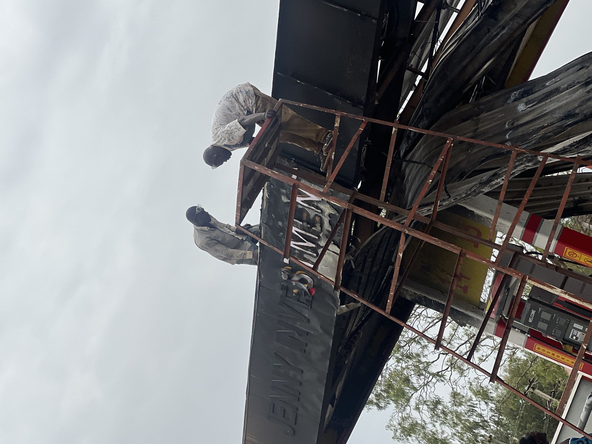 Two individuals are perched on a damaged metal structure, possibly a sign. The word "JENNYMEN" is partially visible on the structure. The individuals are wearing light-colored clothing. The background includes a cloudy sky, trees, and a building. The scene appears to be in Gura Top, Nigeria, based on the sign on the gas pump that says Nikrow Oil & Gas.