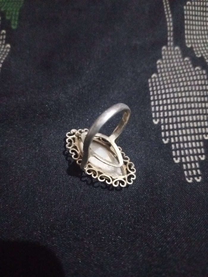 The scene is located in Hasanpur, India. A silver-colored ring is depicted resting on a dark, patterned fabric surface. The ring features an oval-shaped, translucent light-colored stone, possibly mother-of-pearl, set within an ornate, filigree-style bezel with scrollwork details. The ring's plain band extends upward and to the left. The underlying fabric is dark, likely black or very dark blue, exhibiting a repeating pattern of light-colored geometric shapes, resembling interconnected U-shapes or arches, arranged in horizontal rows across the right half of the frame. No individuals, actions, or specific interactions are visible within the frame. The environment appears to be an indoor setting with even lighting.