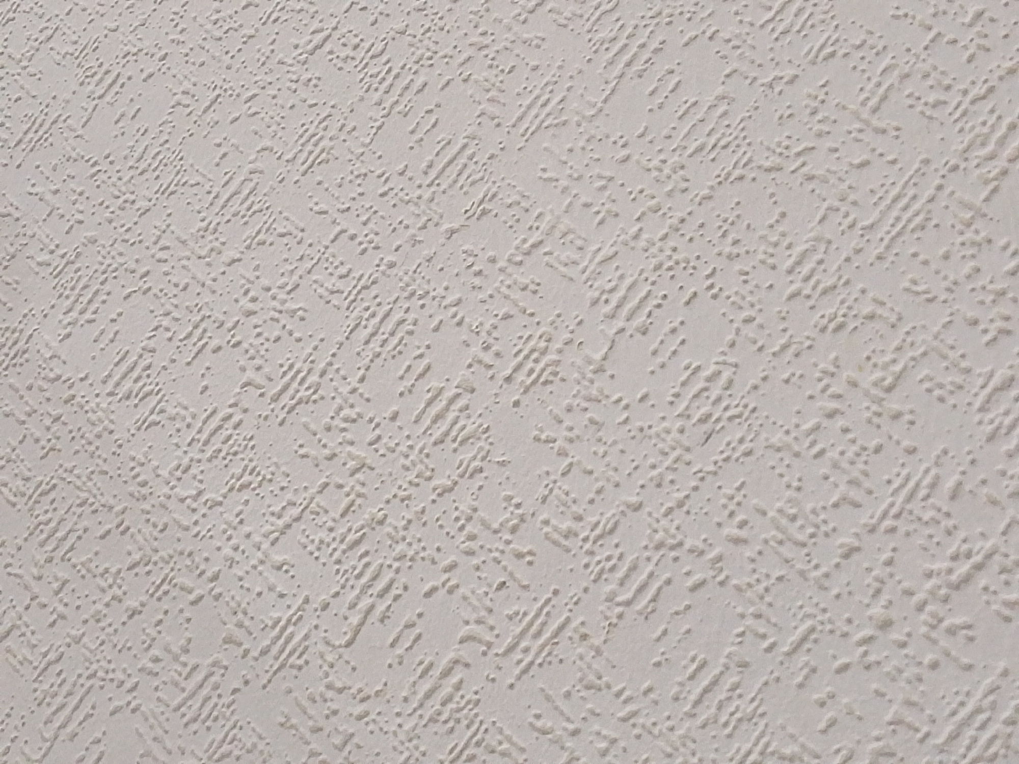 The image shows a close-up, angled view of a textured, off-white surface, likely wallpaper or a painted wall. The texture is characterized by a pattern of raised, irregular lines and dots, creating a slightly bumpy and uneven surface. The lighting casts subtle shadows, highlighting the texture. There are no people, animals, or discernible objects in the frame, nor any indication of a specific urban environment or location within Koltushi, Russia. The image is purely focused on the abstract texture of the surface.