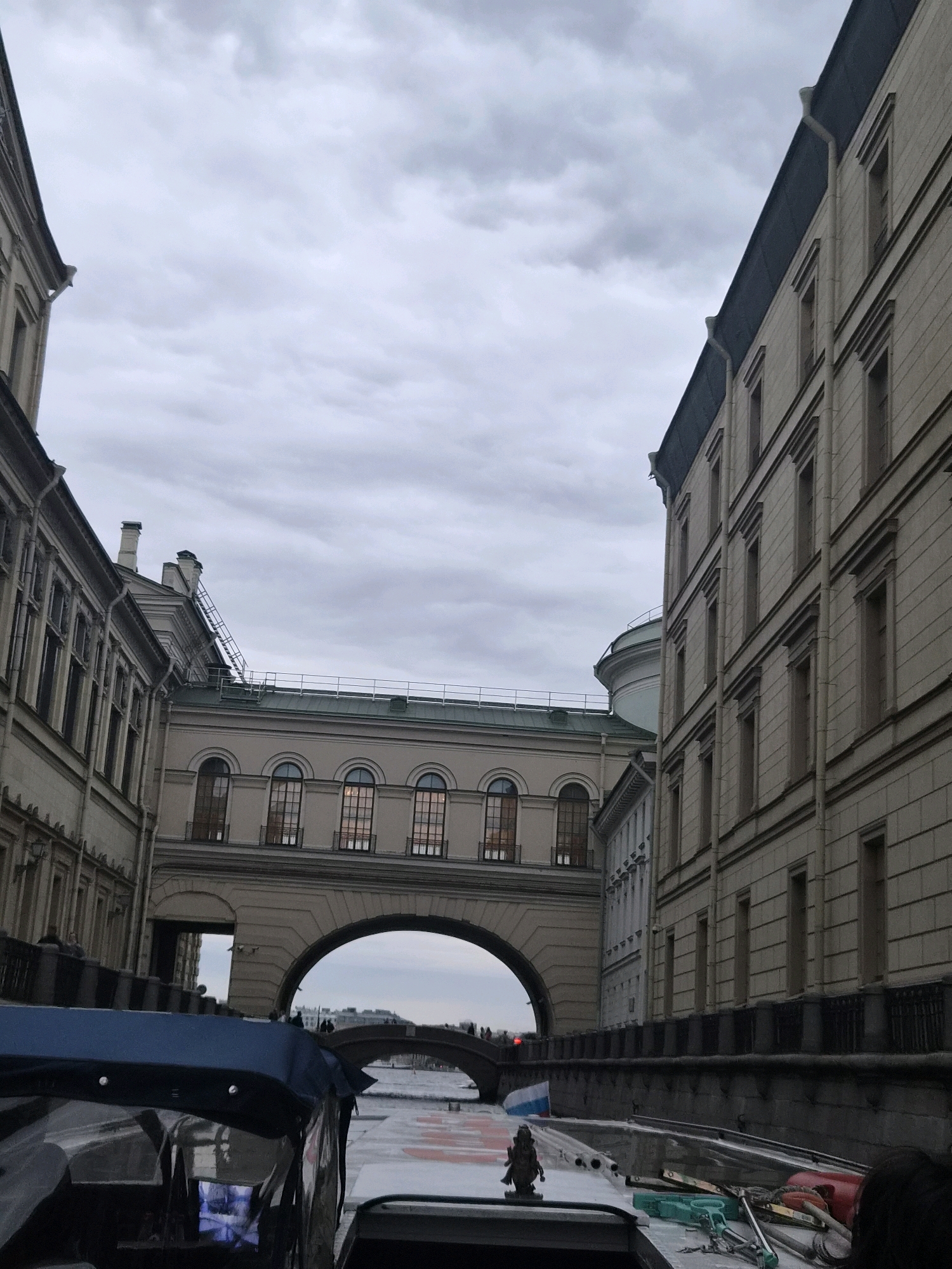 The image captures a canal in Saint Petersburg, Russia, likely taken from a boat. The scene is dominated by grand, classical architecture on both sides of the canal, characterized by pale stone facades, numerous windows, and decorative cornices. A notable architectural feature is a large, arched bridge-like structure spanning the canal, connecting the buildings. The sky is overcast with heavy clouds, casting a subdued light on the scene. The foreground shows the edge of a boat with a blue canopy, some equipment, and a person's head visible. In the distance, another bridge can be seen, along with more buildings and a hint of a cityscape. The overall impression is one of a historic and elegant urban waterway, typical of Saint Petersburg.