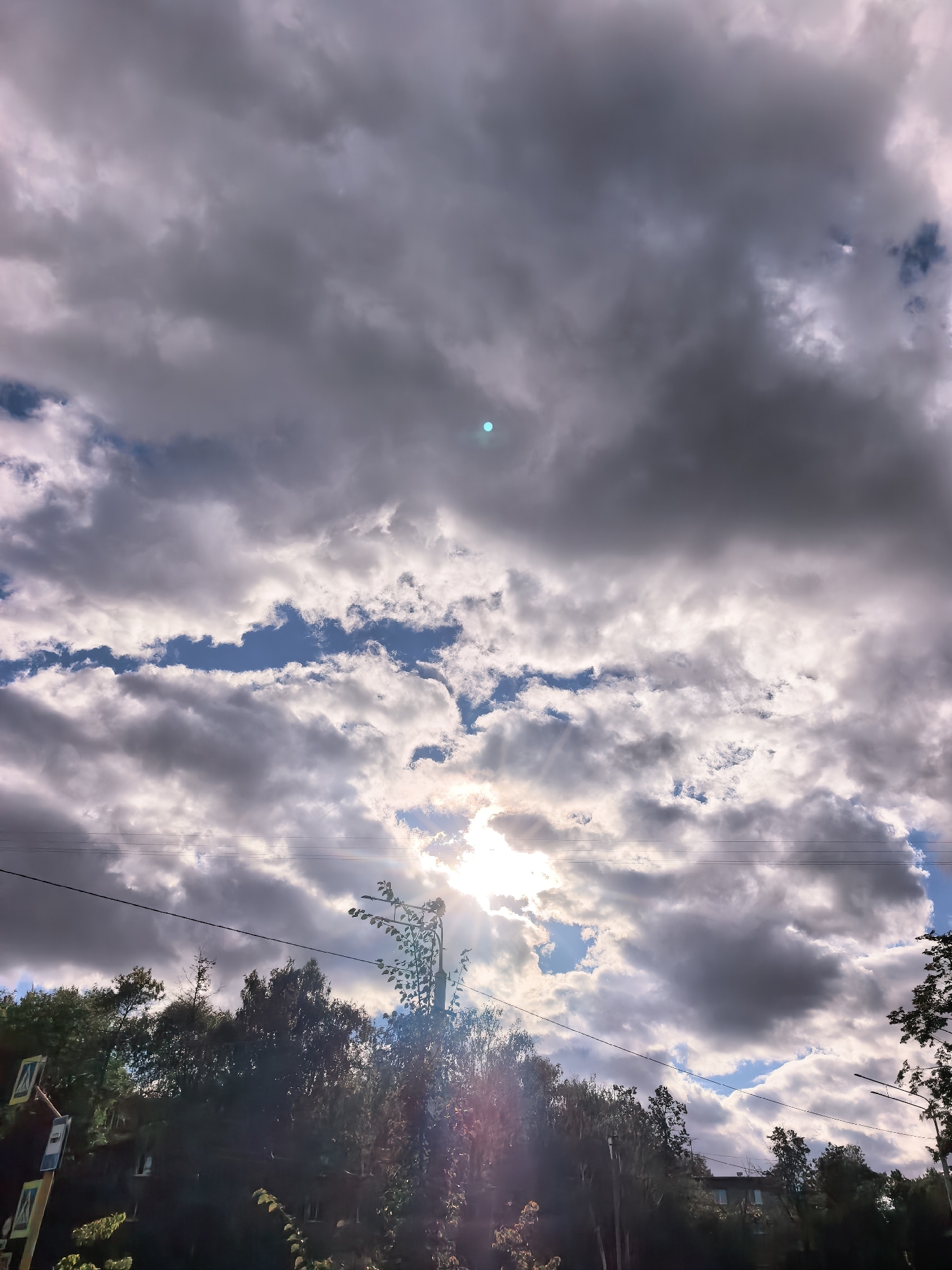 A low-angle, vertical shot captures a dynamic sky over Sterlitamak, Russia, dominated by a dramatic interplay of dark and light clouds. The sun is partially obscured by these clouds, creating rays of light that pierce through, illuminating the scene with a bright, almost ethereal glow. In the lower portion of the image, a silhouette of trees and foliage is visible against the brighter sky. Electrical wires stretch horizontally across the middle ground, adding a man-made element to the natural landscape. A faint, circular blue light is visible in the upper portion of the frame, possibly a lens flare or artifact. The lower section of the image shows hints of buildings and street signs, suggesting an urban or suburban environment.