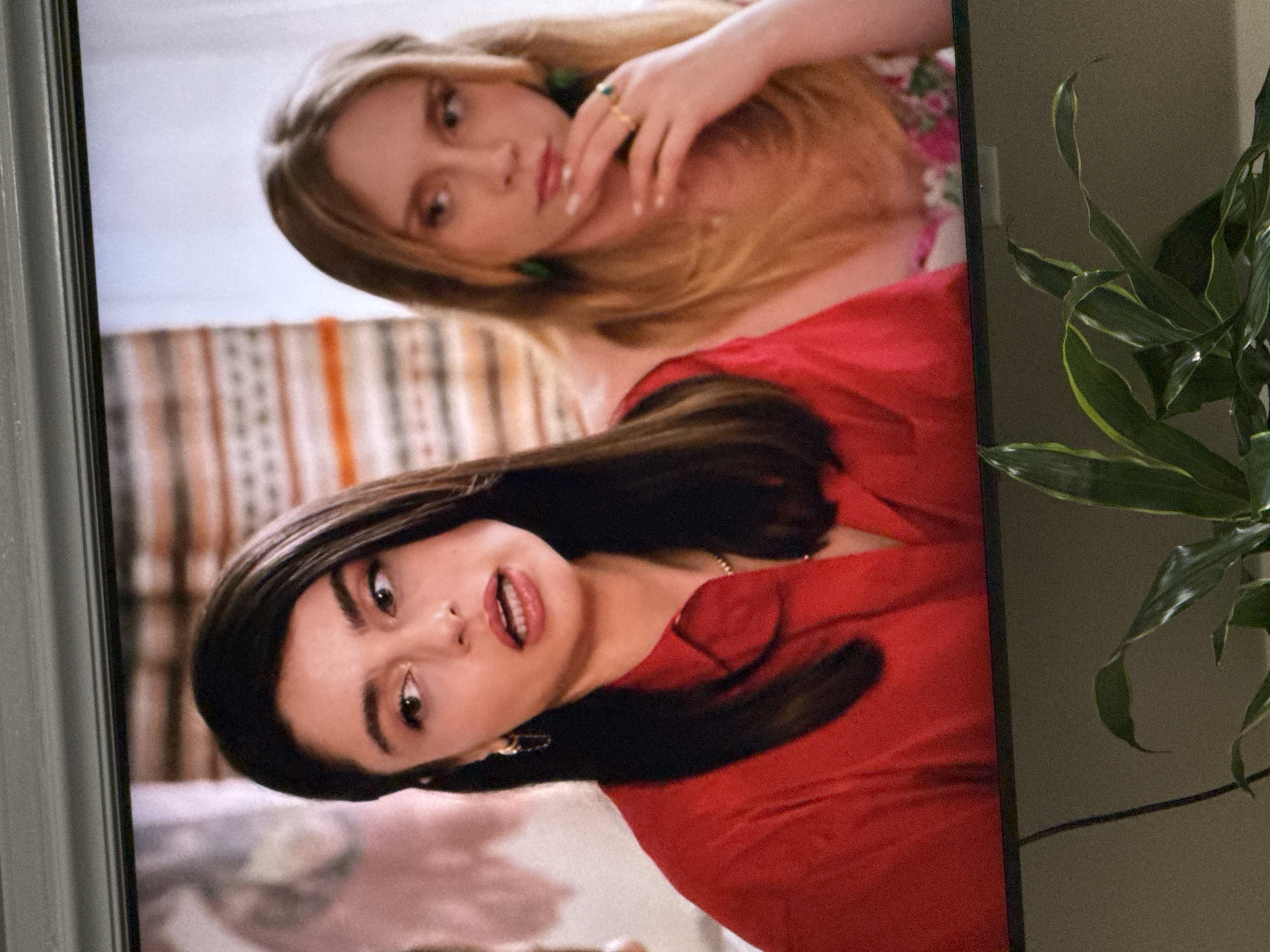 The image shows a close-up of two young women on a television screen. The woman on the left has dark hair and is wearing a red shirt. She is looking to her left with her mouth slightly open. The woman on the right has blonde hair and is wearing a floral top. She is resting her chin on her hand and looking to her left. The background is blurred, but appears to be a room with striped curtains or a tapestry. The bottom of the image shows the top of a television set and a plant with green leaves. The image is taken in the city of Springboro, United States of America.