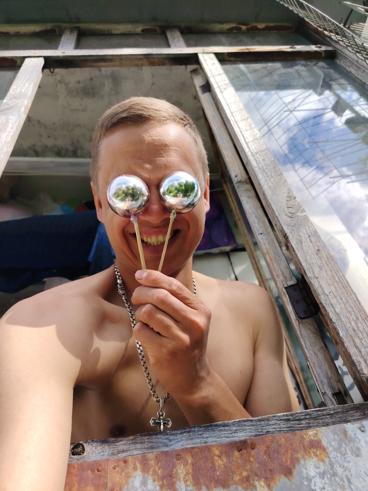 A shirtless man with blond hair and a wide smile is holding two reflective silver spheres in front of his eyes. He is wearing a silver chain with a cross pendant. The man is positioned within an open wooden frame, possibly a window or doorway, with visible signs of rust on the lower part of the structure. Outside the frame, a cloudy sky is reflected in the spheres, and in the background, there are blurred objects and what appears to be a fabric. The overall scene suggests an outdoor or semi-outdoor environment. The location is identified as Chaikovsky, Russia.