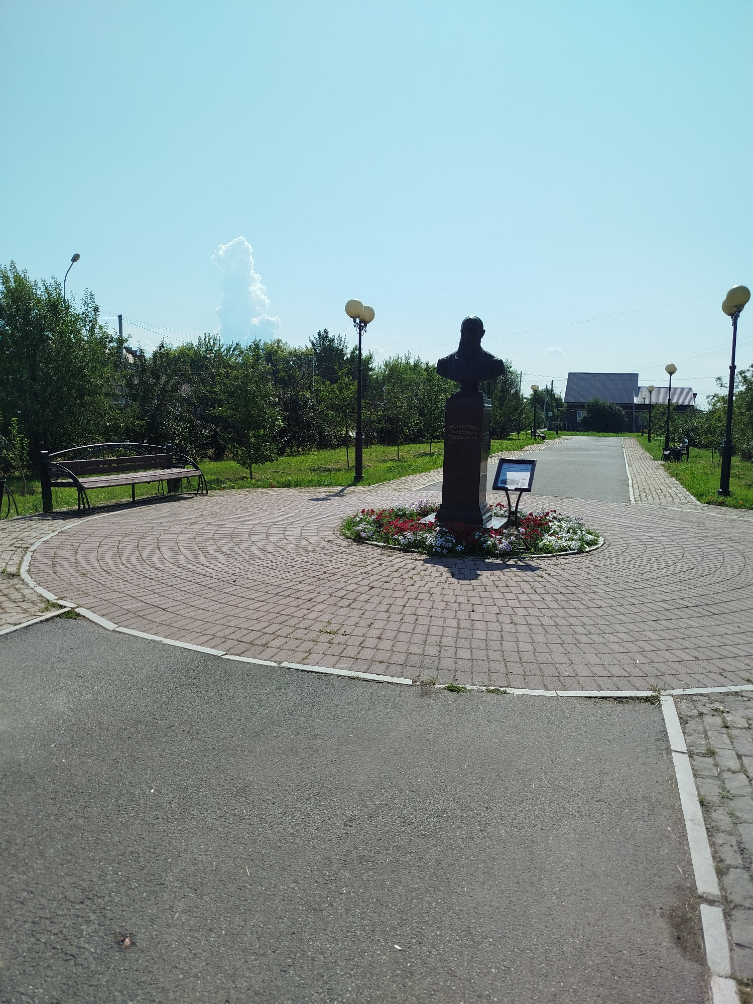 The image depicts a memorial bust in a park located in Yalutorovsk, Russia. The bust, made of dark material, stands on a tall pedestal that is surrounded by a circular flowerbed. The pedestal has an inscription in Russian. A small information board is positioned in front of the bust.
The park features a paved walkway made of red bricks arranged in a concentric pattern around the memorial. To the left of the memorial, a dark-colored park bench is visible. Further back, a road or pathway runs through the park, lined with lampposts. In the background, there are trees, grass, and some houses with dark roofs. The sky is clear and blue with a single white cloud. The scene suggests a quiet, well-maintained public space on a sunny day.