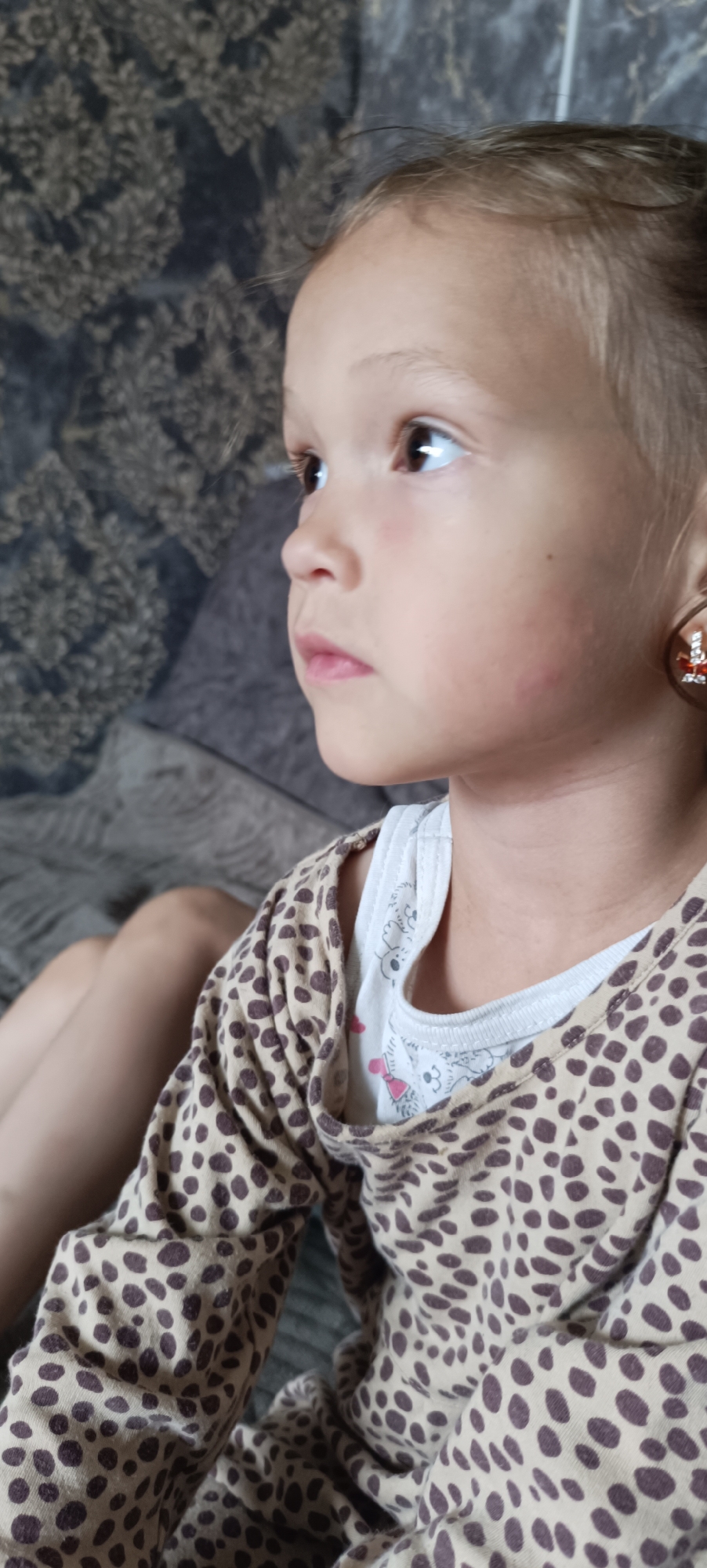 A young girl with light brown hair is seated indoors, looking to her left. She is wearing a light-colored t-shirt with cartoon animal prints and a tan shirt with dark brown leopard spots over it. She has a single hoop earring in her left ear with a red and silver charm. The background consists of a patterned dark fabric couch. The image is a close-up shot, focusing on the girl's face and upper body. There is no indication of the city of Karsha, Russia, in the image.