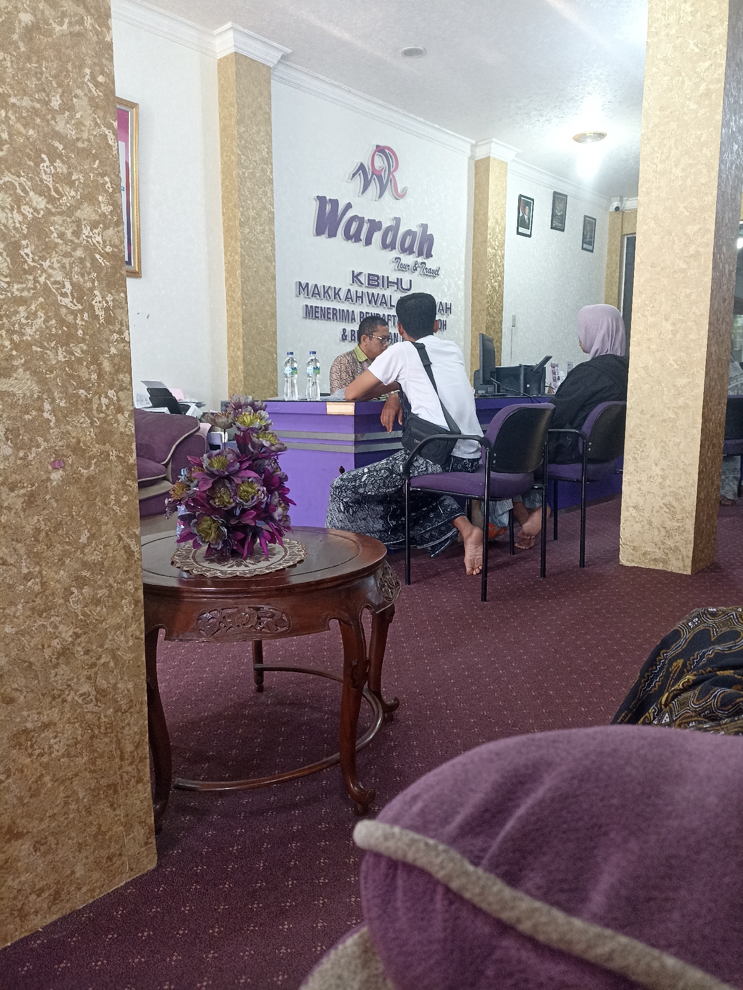 The image shows the interior of a travel agency, likely "Wardah Tour & Travel" based on the signage. The location is identified as Tompokersari, Indonesia. In the foreground, a wooden table with carved details holds an artificial floral arrangement. The carpeted floor is a deep purple. Behind the table, several people are visible. Two men and a woman are seated at a counter. One man, wearing glasses and a patterned shirt, appears to be interacting with the other man, who is wearing a white shirt and a sarong, and has a dark bag slung across his shoulder. A woman, wearing a light purple headscarf and dark clothing, is seated in a purple chair facing away from the camera, also at the counter. Behind the counter, a printer and bottles of water are visible. The walls are decorated with a textured wallpaper and framed pictures. The overall impression is of a business establishment with a waiting area and service counter.