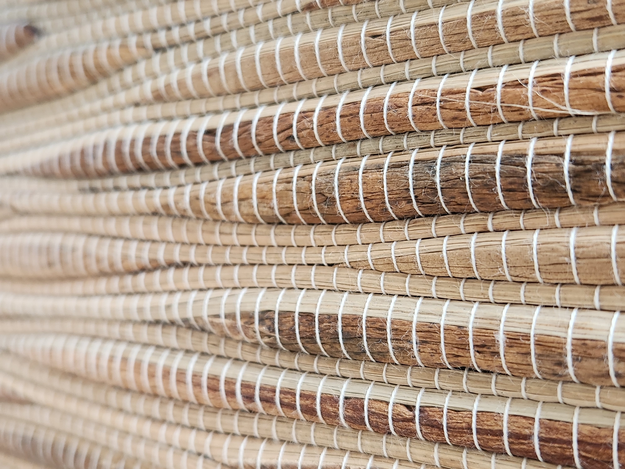 This is a close-up, eye-level, vertical shot of a woven mat. The mat is made of natural fibers, likely straw or reeds, and is predominantly beige and brown in color. The fibers are arranged in vertical rows, with thin white thread wrapped around each row at regular intervals, creating a striped pattern. The texture is rough and organic, with variations in the thickness and color of the fibers. The depth of field is shallow, with the foreground in focus and the background softly blurred. The lighting is natural and diffused, highlighting the texture of the mat. There are no people, animals, or discernible objects other than the mat itself. The image does not provide any specific information about its location in Maitland, United States of America.