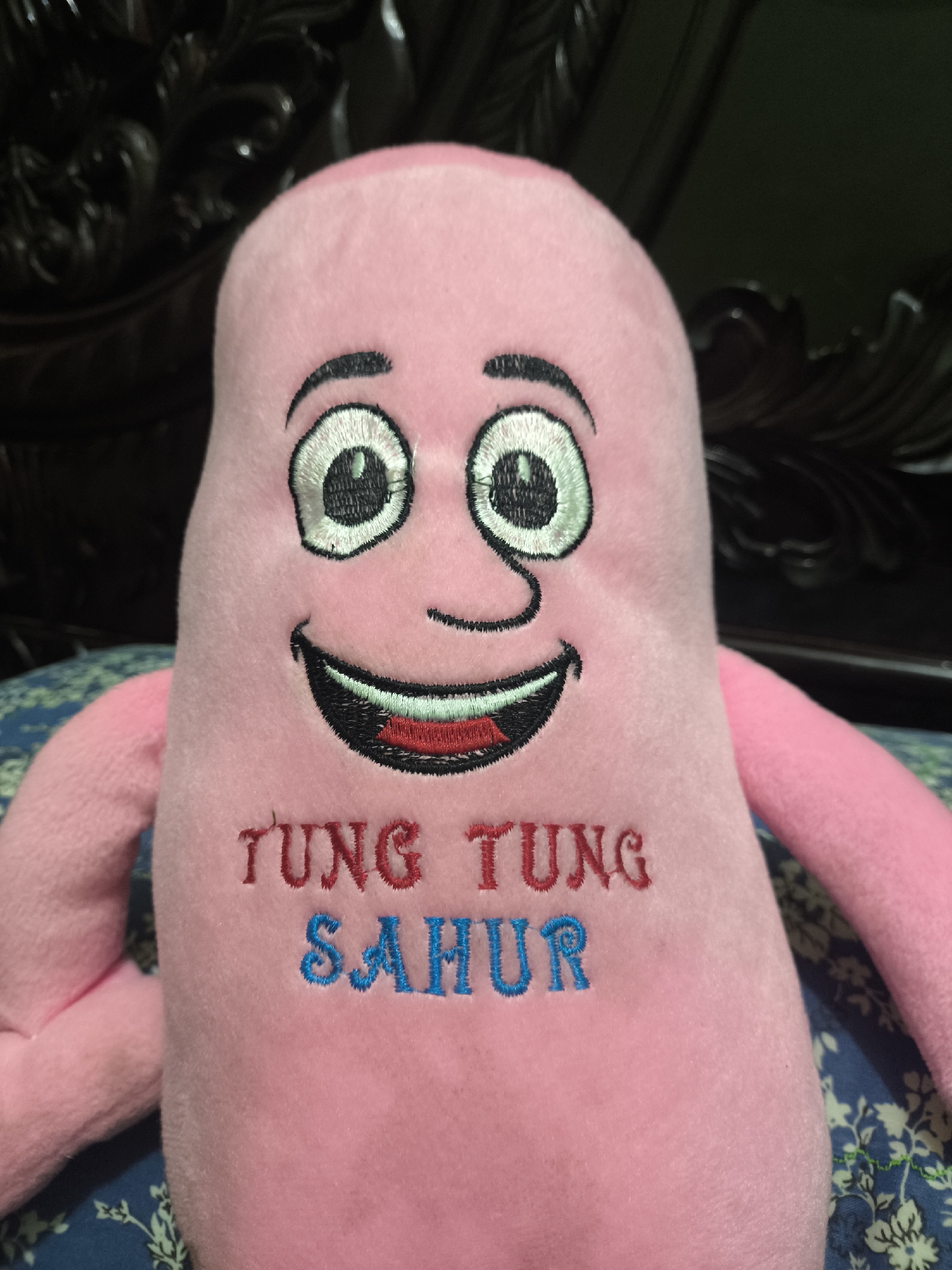 The image shows a close-up of a pink plush toy with a smiling face. The toy has embroidered features, including large eyes with black outlines and white and green centers, eyebrows, a nose, and a wide mouth with red lips and black teeth. The words "TUNG TUNG SAHUR" are embroidered on the front of the toy in red and blue lettering. The toy is positioned against a dark, ornate background, possibly furniture, and rests on a blue and green patterned fabric. The context provided by "Ujongreuba, Indonesia" suggests the image was taken in that location.