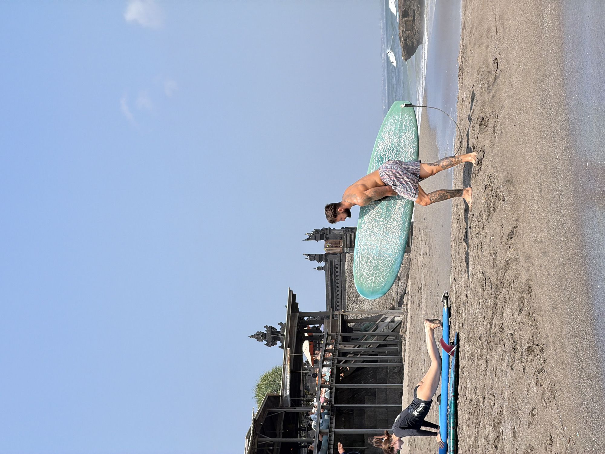 In North Kuta, Indonesia, on a sandy beach, a shirtless man with tattoos carries a large, light blue surfboard. He is wearing patterned swim trunks and walking towards the right of the frame. In the foreground, a woman wearing a black rashguard and black bikini bottoms is on the sand in a plank position on top of a blue surfboard. Behind them, a multi-level wooden building with a thatched roof sits on a raised stone foundation. The building has people seated on its decks, some with umbrellas. The ocean is visible in the background with gentle waves and a rocky outcrop. The sky is clear and blue.