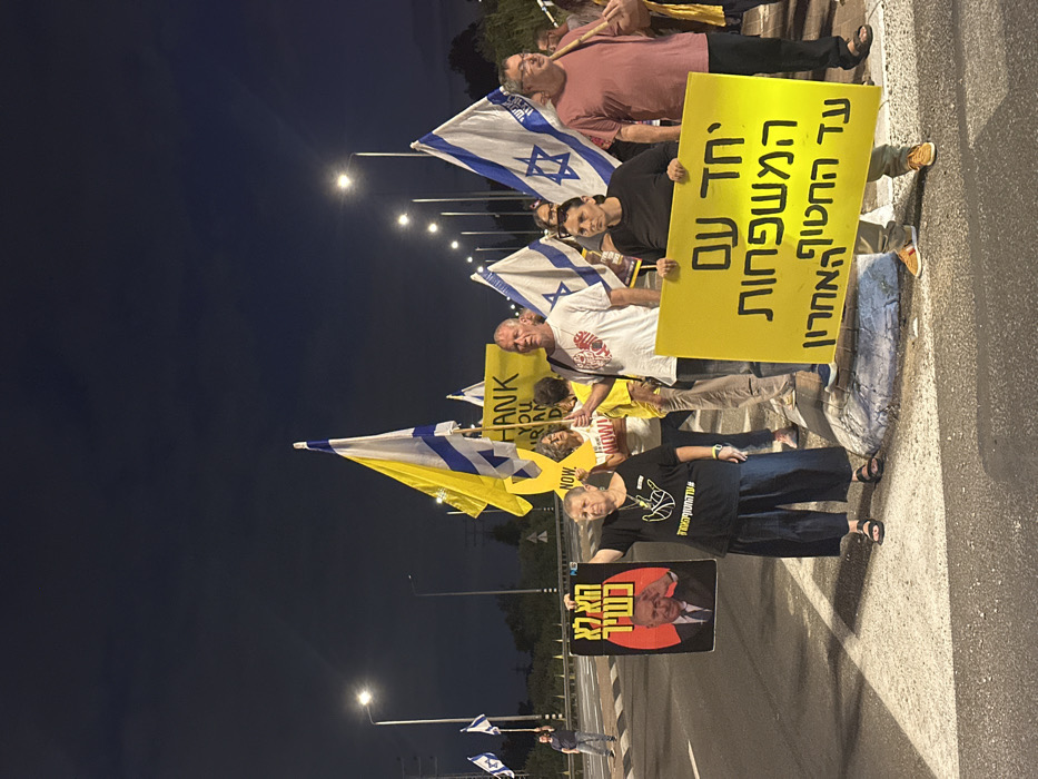 At night in Mishmar Hasharon, Israel, a group of approximately 10-15 individuals is engaged in a stationary protest on an asphalt road with white markings, adjacent to a raised concrete median. The scene is illuminated by multiple streetlights.
Prominent in the foreground are three individuals. On the left, a woman with short, light hair, wearing a black t-shirt and dark pants, holds a black rectangular sign featuring a portrait of Benjamin Netanyahu. Red Hebrew text on the sign reads "הוא לא כשיר" (He is not fit). To her right and slightly behind, a man in a white t-shirt with a red design holds an Israeli flag. In the center foreground, another woman in a black top and light pants holds a large, rectangular yellow sign with Hebrew text that reads "יחד עם המשפחות עד החטוף האחרון" (Together with the families until the last hostage). To her right, a man in a reddish-brown t-shirt, glasses, also holds an Israeli flag.
Several other individuals are visible in the mid-ground and background, holding additional Israeli flags, some combined with yellow fabric, and other yellow protest signs, one partially visible with English text "THANK YOU NOW!". A man in the far left background also holds an Israeli flag.
The environment includes a multi-lane road, a white guardrail in the mid-ground, and numerous streetlights extending into the distance under a dark night sky. Power lines are visible on the left side.