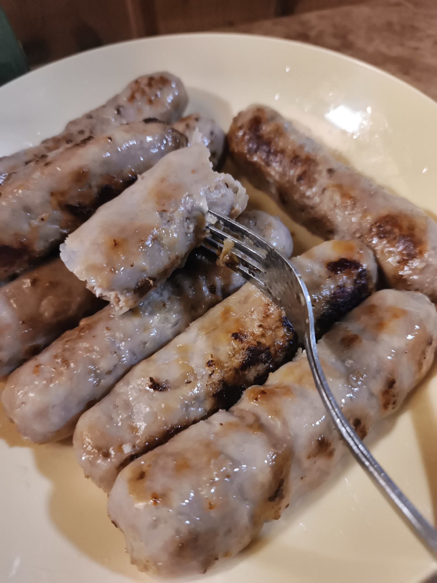 A close-up shot captures a plate of cooked sausages on a light yellow plate. A fork is shown in the center, with its tines piercing one of the sausages, and slightly lifting it to reveal its interior. The sausages are plump and have a slightly browned, glistening appearance, indicating they are likely grilled or fried. Some sausages show darker, slightly charred spots. The background is blurred, showing hints of brown and dark colors, possibly a table or wooden surface. The lighting creates highlights on the greasy surface of the sausages.