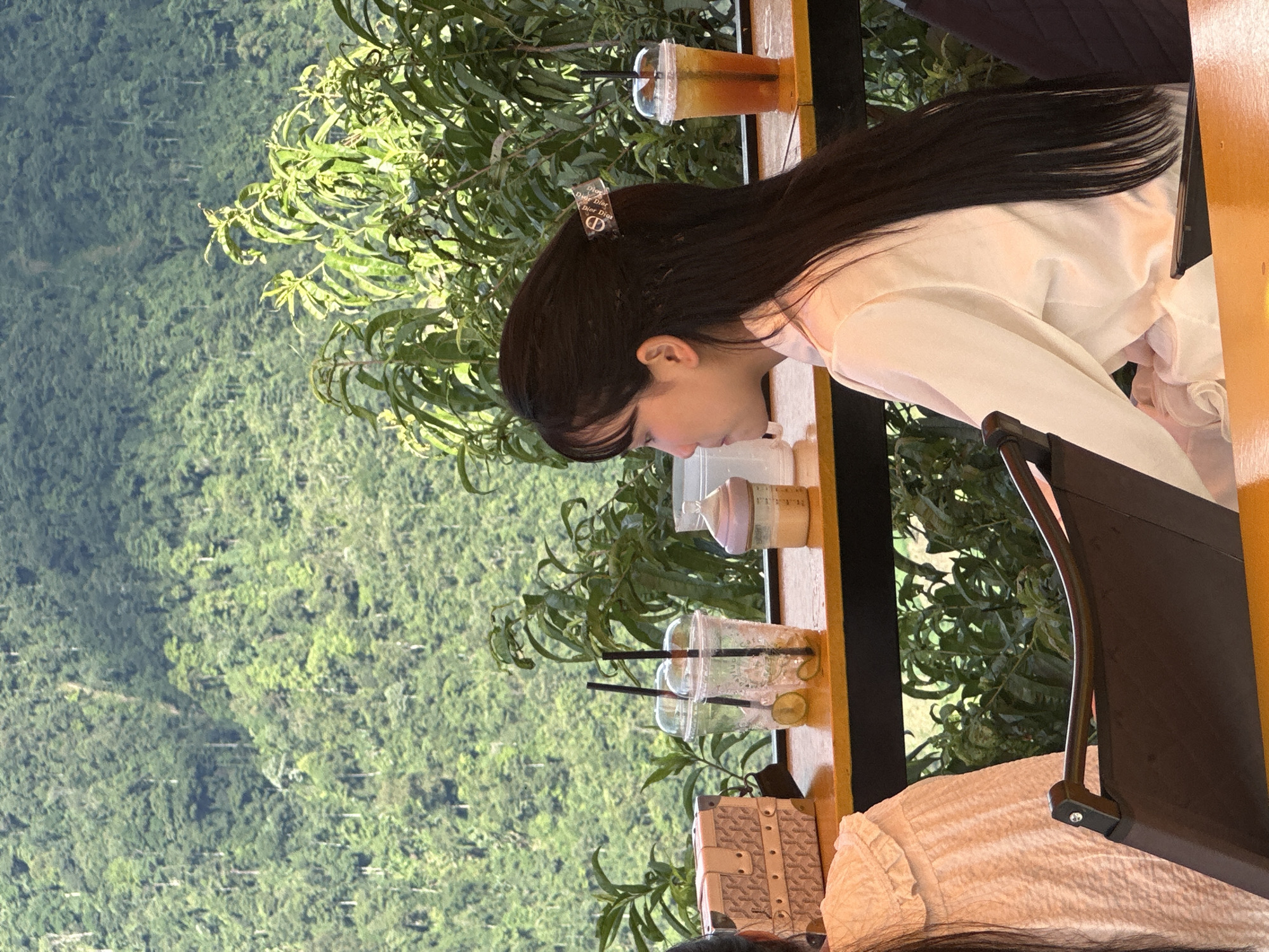 The image captures an outdoor cafe scene in Tu Le, Vietnam, presenting a side profile view of a young woman seated at a wooden counter. She has long, straight dark hair, partially held back by a clear hair clip featuring a repeated "Dior" pattern/logo. She is wearing a light-colored, long-sleeved top and is looking to her right, away from the camera. To her immediate left, the back of a second person is partially visible, wearing a light-colored top with ruffles.
On the wooden counter in front of the individuals, from left to right, are several items: a light brown rectangular box with a repeating geometric pattern; two clear plastic cups with lids, black straws, ice, and lime slices (one mostly empty, the other partially full); a small, light pink/purple bottle with a nipple-like cap containing a light brown liquid; and a clear plastic cup with a lid and a black straw, containing an amber-colored iced beverage with ice, which appears mostly full.
Immediately behind the counter is a row of lush green foliage and tree branches. Beyond this midground vegetation, the background is dominated by a vast, dense, and verdant mountain range covered in abundant green trees, illuminated by natural daylight.