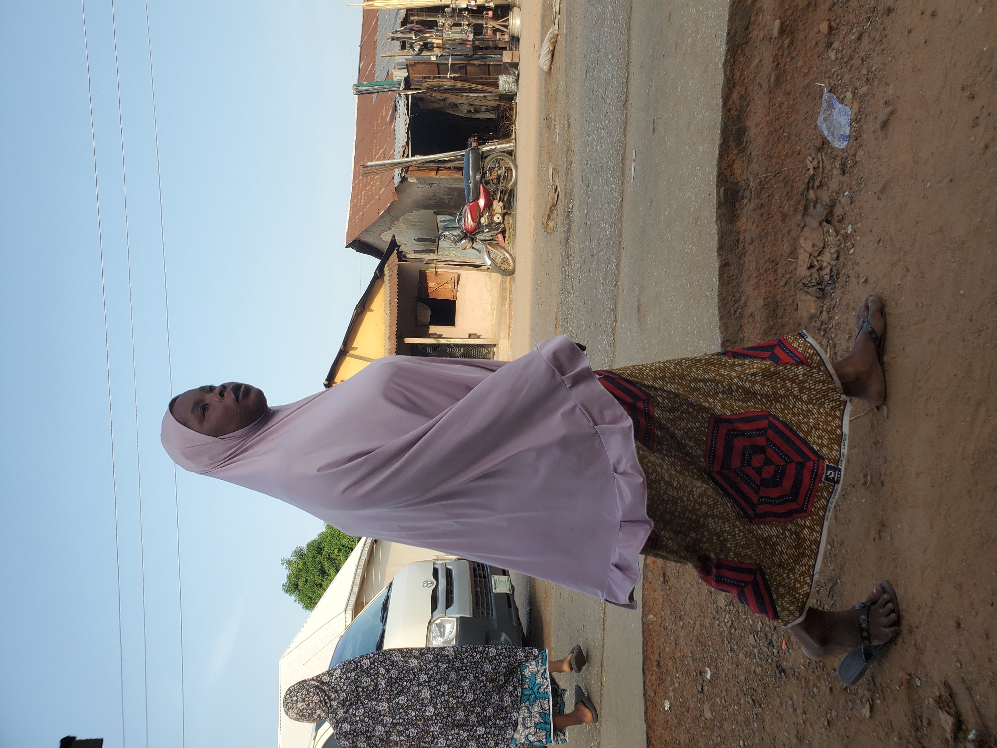 A woman wearing a light pink hijab and a long, patterned dress is walking along a street in Mararaba, Nigeria. She is on the right side of the road, which is partially paved with a strip of exposed dirt to the right. She is wearing open sandals. Another woman in a floral dress and hijab stands beside a white van. Across the street are corrugated metal buildings, one of which houses a motorbike. Plastic litter is scattered on the ground. The sky is light blue.