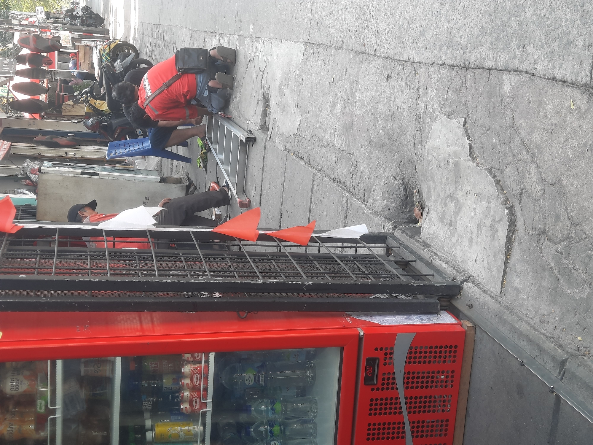 In North Jakarta, Indonesia, a street scene unfolds where two individuals are observed interacting with a ladder positioned on a cracked concrete sidewalk, adjacent to a red beverage cooler. One person, wearing a red shirt, kneels, while another, in dark pants and a cap, stands. The cooler, branded with a Coca-Cola logo, is filled with bottled drinks. In the background, parked motorcycles and a blue plastic chair suggest a commercial area. The environment is an urban setting with pavement and buildings. The lighting suggests it is daytime.