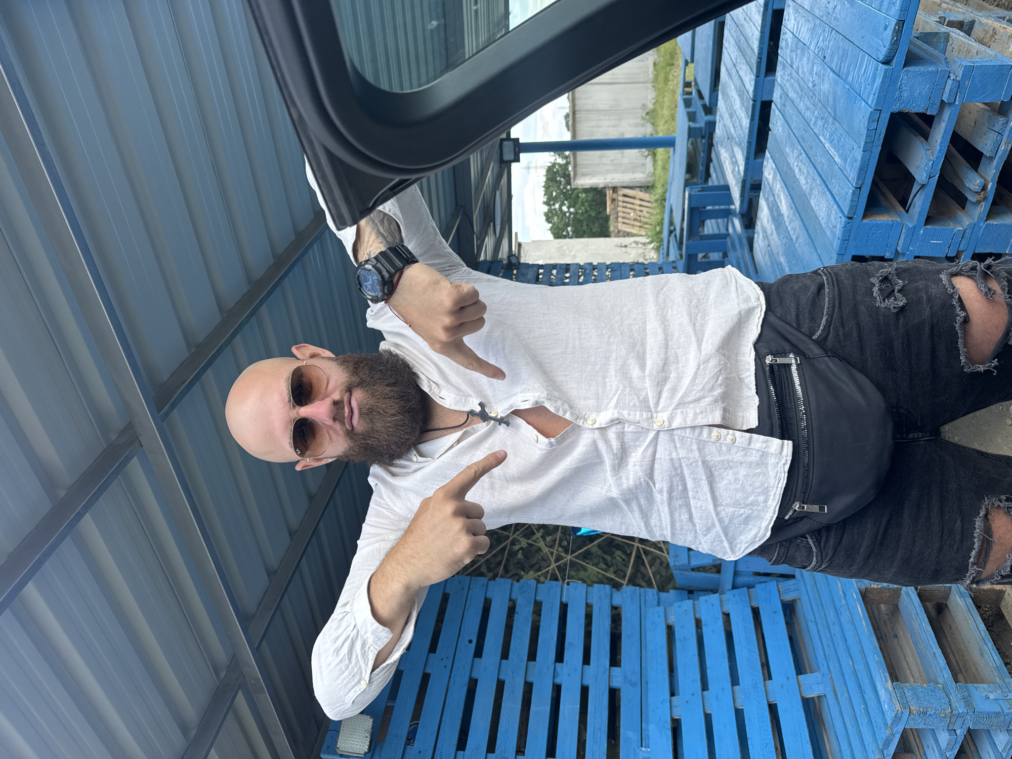 A bald man with a beard is standing under a metal structure. He is wearing sunglasses, a white shirt, ripped black jeans, and a black fanny pack. He is pointing down with both hands. The background includes blue wooden pallets and a glimpse of a building with trees.