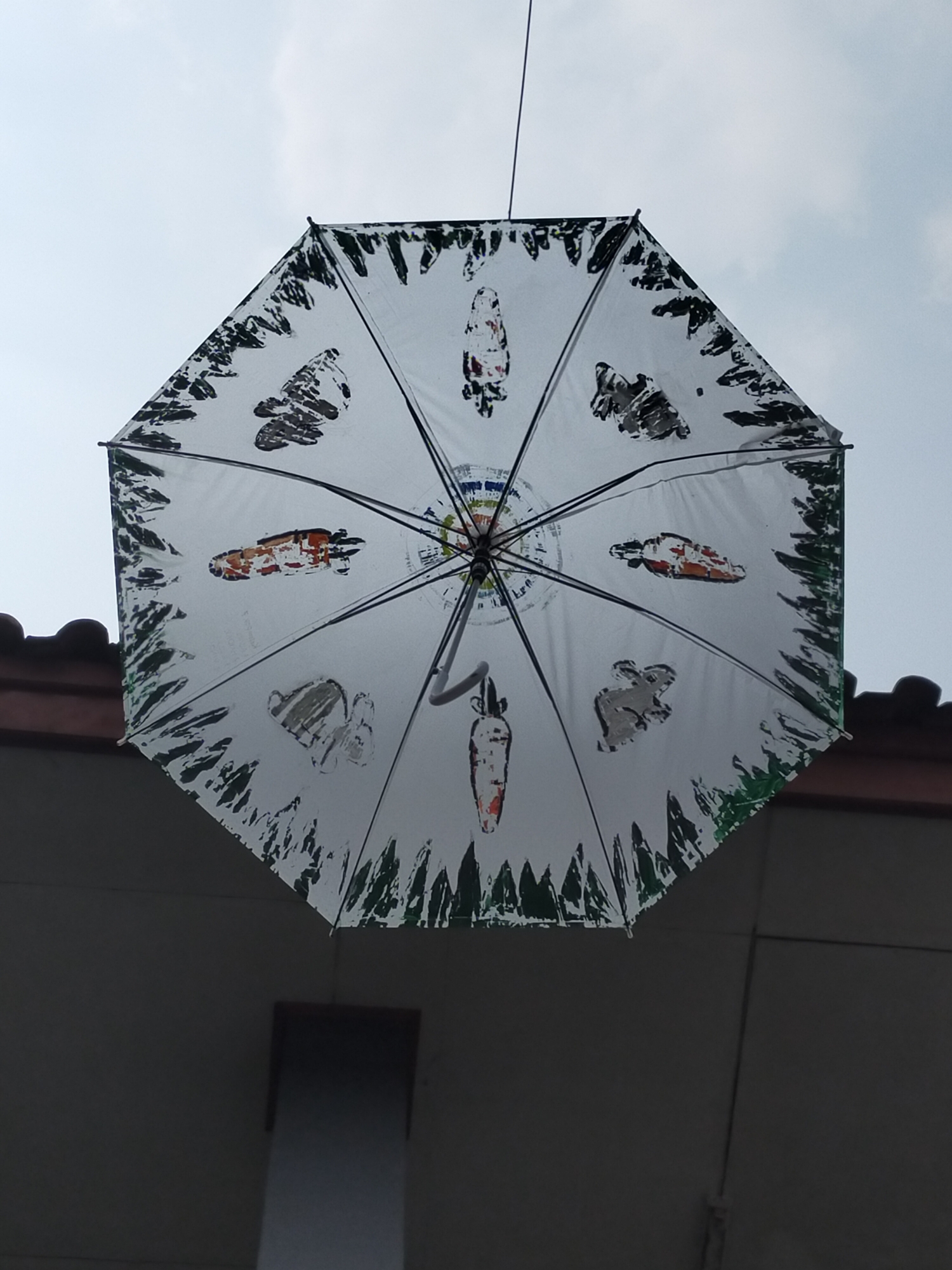 An octagonal white umbrella decorated with dark, abstract designs and a few colorful images, including carrots and what appears to be a rabbit, is suspended in the air against a cloudy sky. The umbrella is positioned above the tiled roof of a building. Below the roof, the side of a building is visible in shadow, with a rectangular object hanging from it. This scene is located in Tambun, Indonesia.