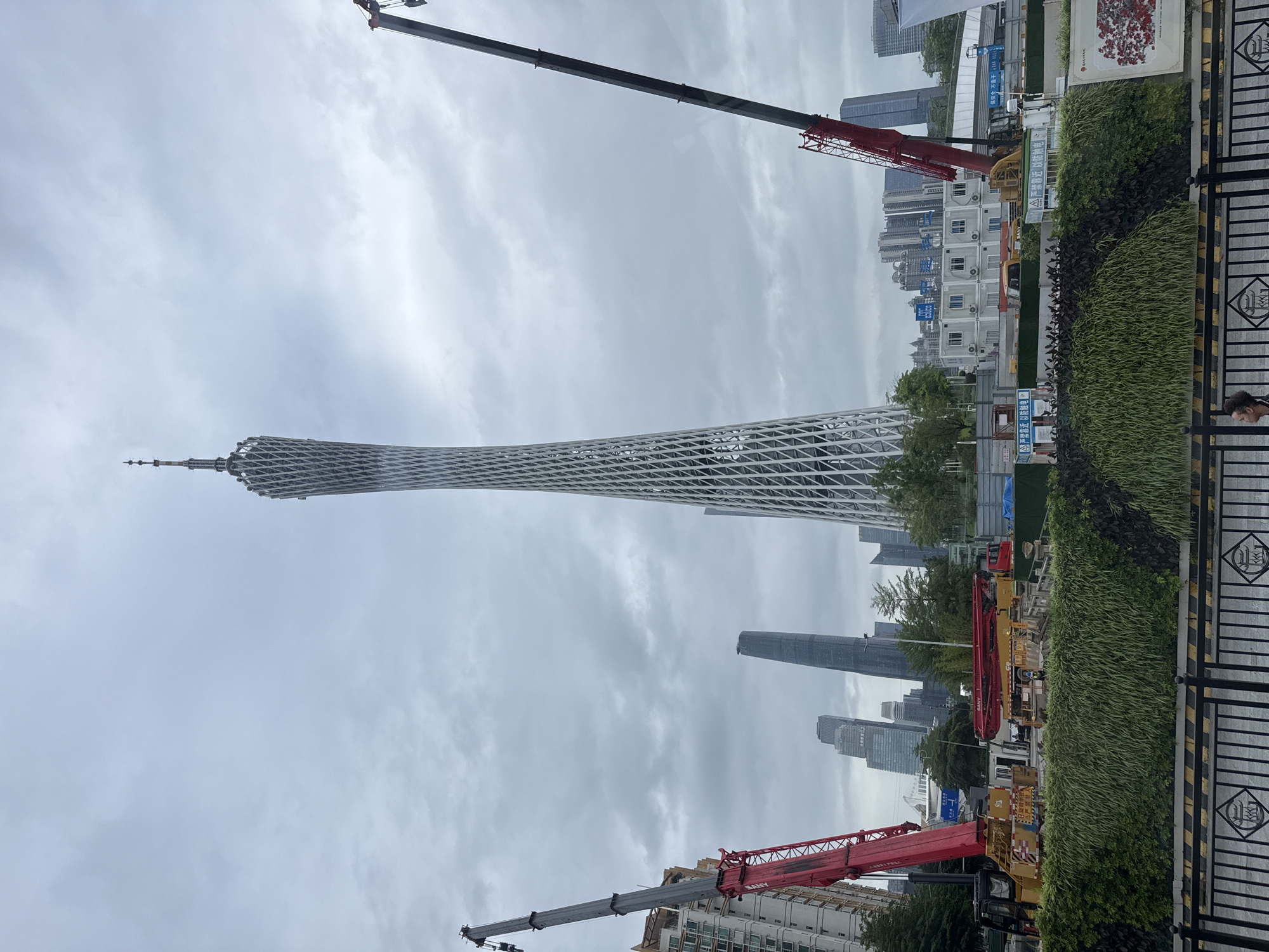 The image depicts the Canton Tower in the Haizhu District of Guangzhou, China, under an overcast sky. The tower is prominently featured, along with several other high-rise buildings in the background. Two construction cranes with red booms and grey jibs are visible, one in the upper left and the other in the lower part of the frame. A green hedge borders the foreground, with sections of a black metal fence visible. In the background are several portable construction offices. The overall scene suggests a construction site or urban development area within the city. A person's head is visible in the lower right corner, behind the hedge.