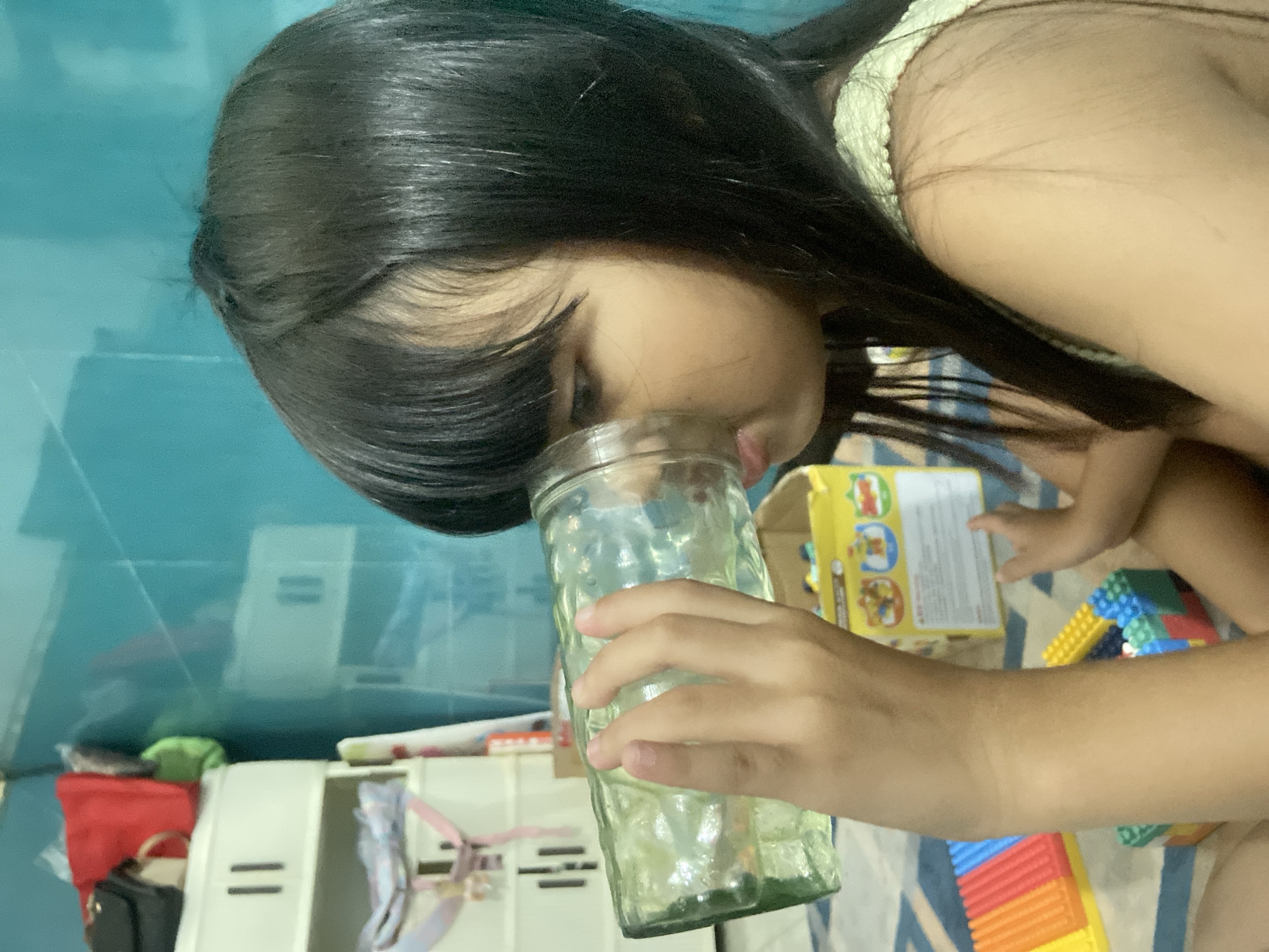 A young girl is sitting on the floor, holding a clear, textured glass to her face, appearing to drink from it. She has dark hair and is wearing a light green top. The background features a turquoise wall with shelving units, some with items on them, including bags and what appears to be a toy box. Colorful building blocks are scattered on the floor around her, suggesting a playroom setting. The scene is indoors, with the lighting indicating it's daytime. There is no indication that this location is in Gelam, Indonesia.