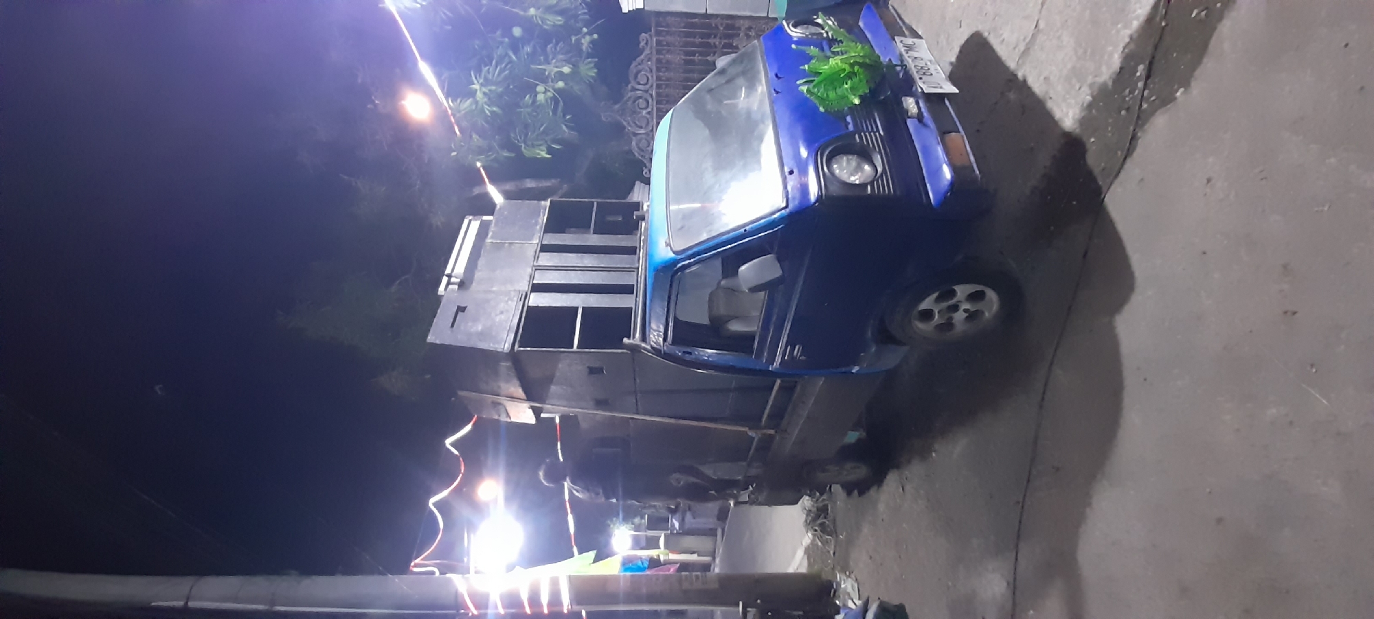 In Iser, Indonesia, a blue pickup truck is parked on a paved surface, likely a street, at night. The truck is heavily modified with a large, multi-tiered speaker system loaded into the bed and stacked high. The speakers appear to be enclosed in dark, rectangular cabinets. One person is visible standing on the rear of the truck, near the speakers, facing away from the camera. Decorative lights, possibly for an event, are strung across the background, casting a warm glow. The truck's headlights are on, and a cluster of green foliage is affixed to the front bumper. A license plate is partially visible on the front of the truck. The overall scene suggests a mobile sound system setup, possibly for an outdoor event or celebration.