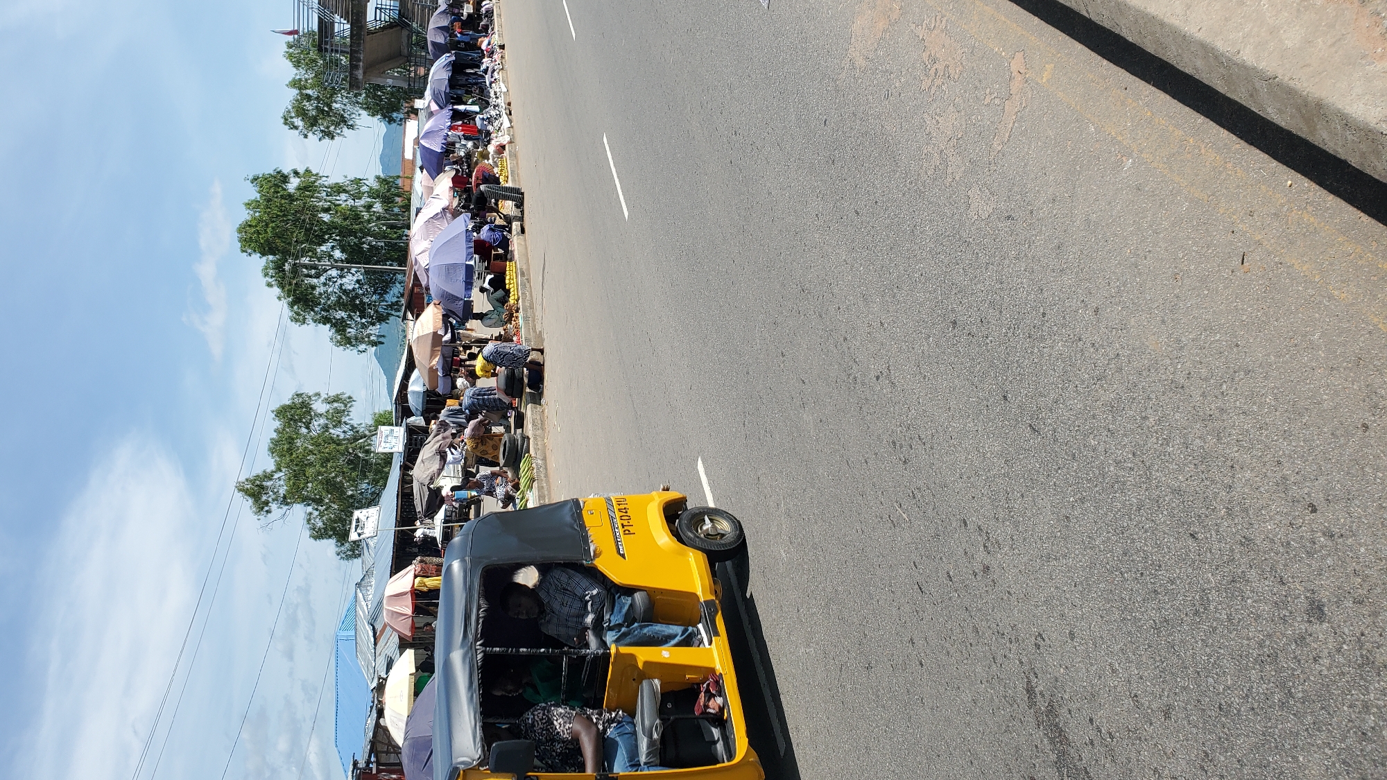 The image depicts a busy street scene in Jalingo, Nigeria, likely a marketplace. In the foreground, a yellow auto-rickshaw, also known as a "Keke Napep" or "Maruwa," is visible, carrying passengers. The rickshaw has the registration number "PT-0410" displayed. Along the side of the road, numerous market stalls are set up under various umbrellas, displaying a range of goods. People are engaged in buying and selling activities. The environment appears to be a public space with a paved road and a sidewalk area where the market is situated. Tall green trees are present in the background, and the sky is partly cloudy. The overall impression is one of a vibrant, active urban market.