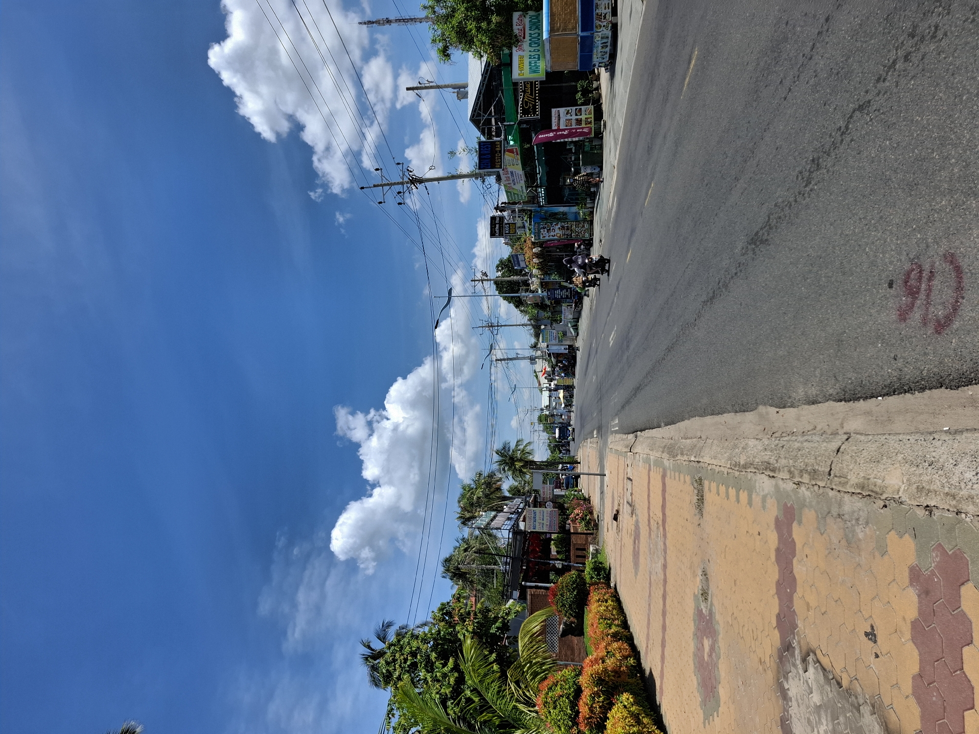 This is a ground-level, wide-angle shot of a street in Phu Thuy Ward, Vietnam, on a sunny day with a blue sky dotted with white clouds. The scene depicts a road with two lanes of traffic. On the left side of the road, there is a sidewalk paved with yellow and red interlocking bricks. A manicured hedge of orange and green bushes lines the edge of the sidewalk. Beyond the hedge, there are palm trees and buildings with storefronts and signs, suggesting a commercial area. On the right side of the road, more storefronts are visible, some with green awnings. Electrical poles and wires run along the street. In the middle of the road, a few motorcycles are visible, one with two riders. In the distance, there appears to be a bus or truck. The overall impression is a bustling street scene in a tropical or subtropical climate.